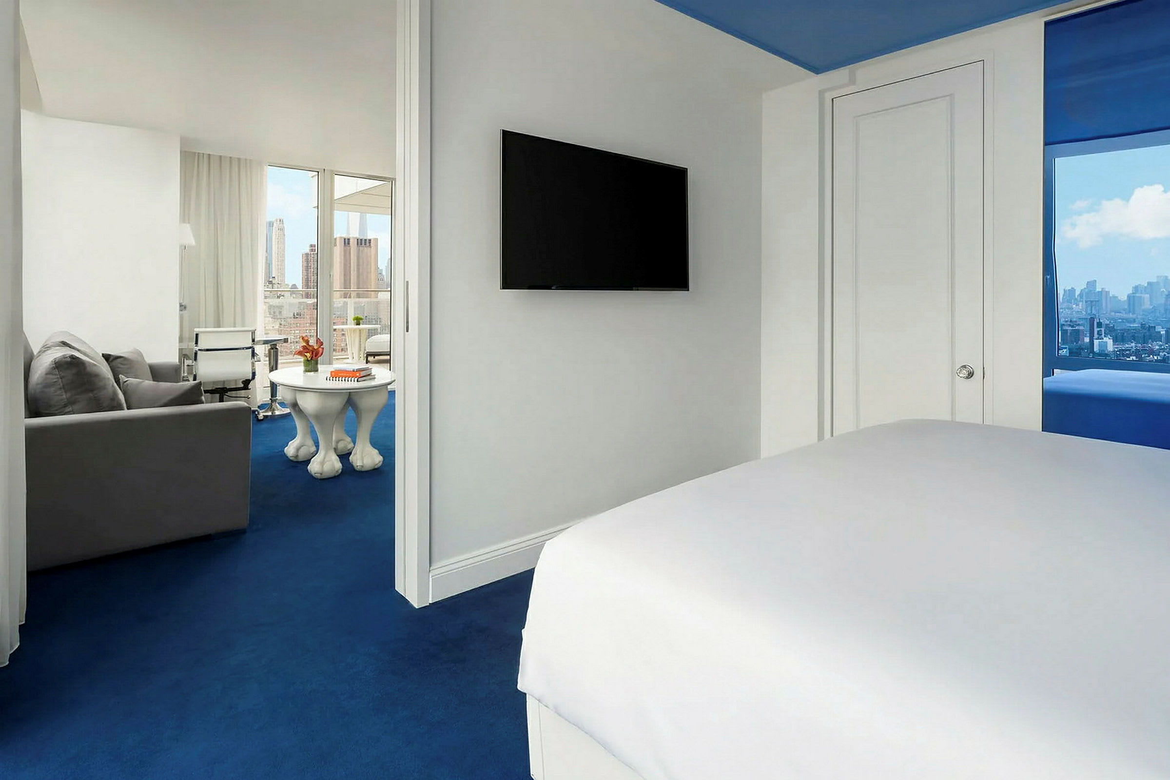 Hotellrom med hvit innredning og blått teppe. Sengen kan sees delvis i forgrunnen, en TV henger på veggen, og i bakgrunnen er det en sittegruppe med utsikt over byen. (AI-generert tekst)