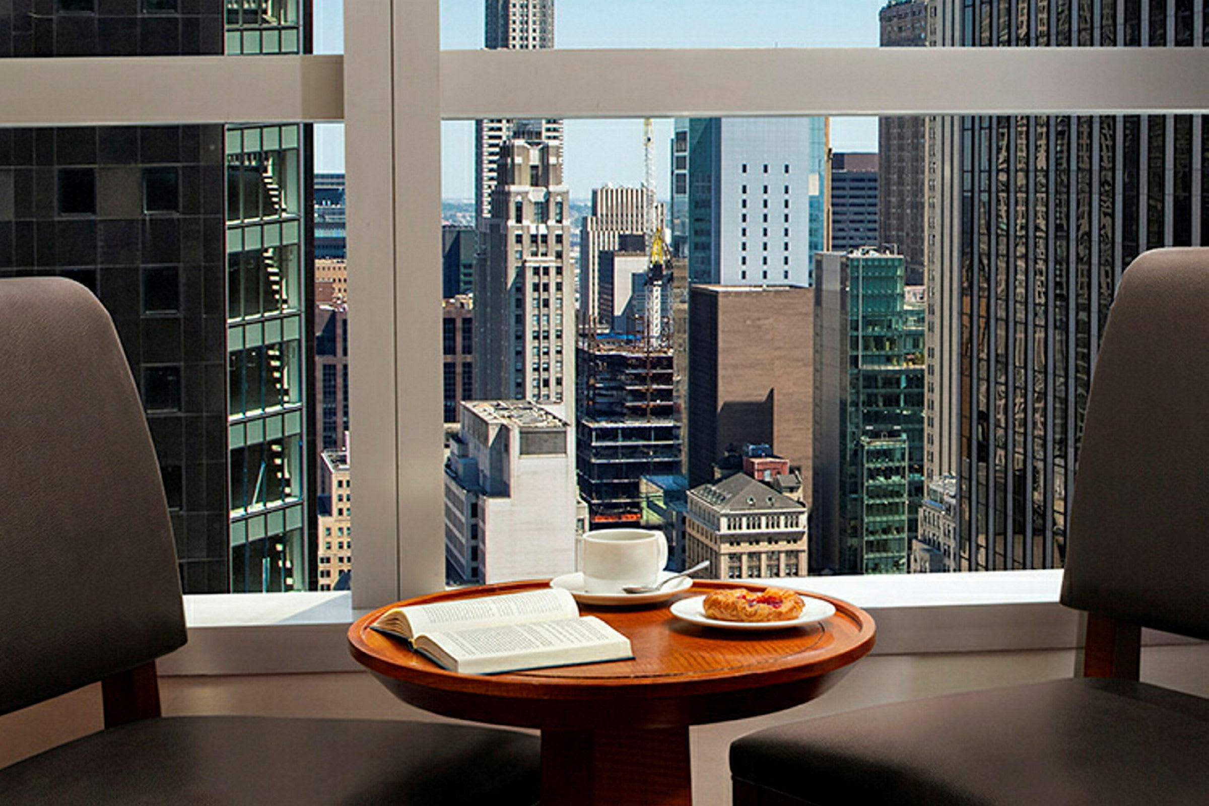To stoler ved et bord med en kopp kaffe, en tallerken med bakverk og en åpen bok, med byutsikt gjennom et vindu. (AI-generert tekst)