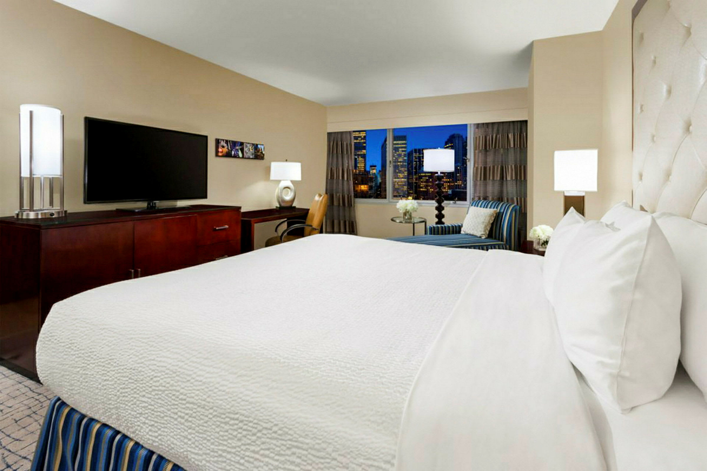 Et smakfullt innredet hotellrom med en dobbeltseng, en TV på en kommode og et sitteområde ved et vindu med utsikt over byens skyline om natten. (AI-generert tekst)