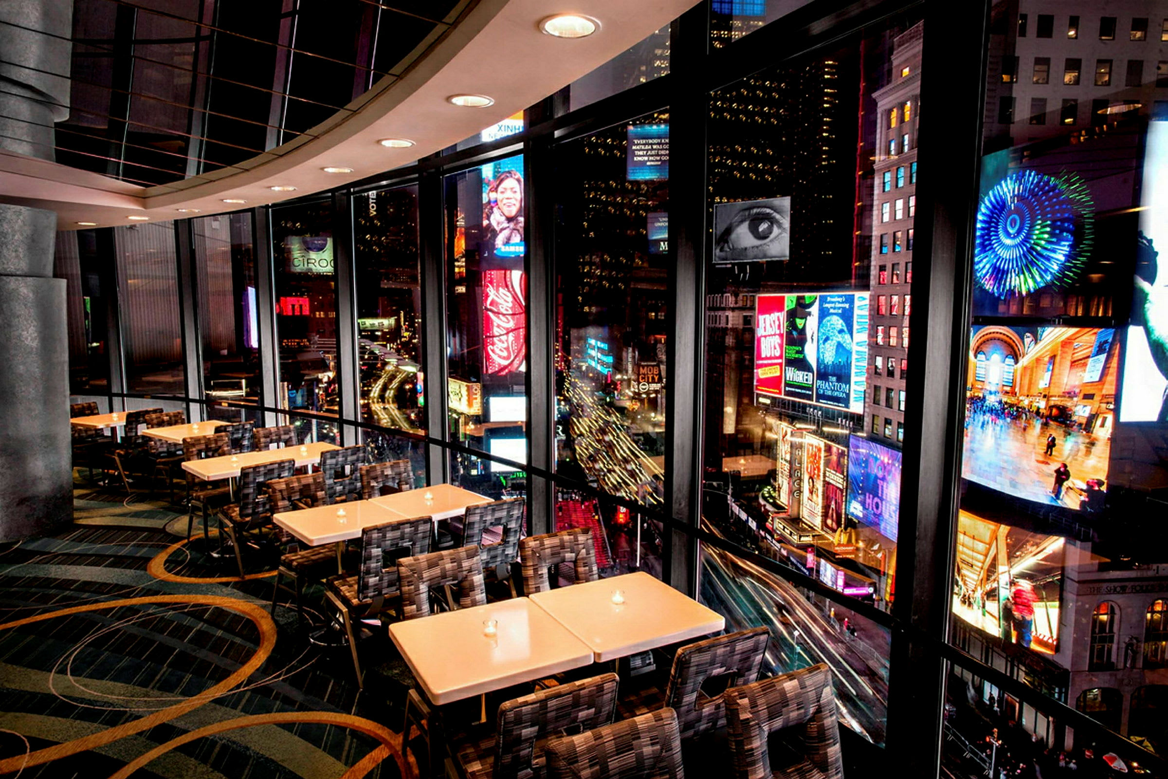 En restaurant med moderne interiør og store panoramavinduer som vender mot lysreklamer og byliv utenfor. (AI-generert tekst)