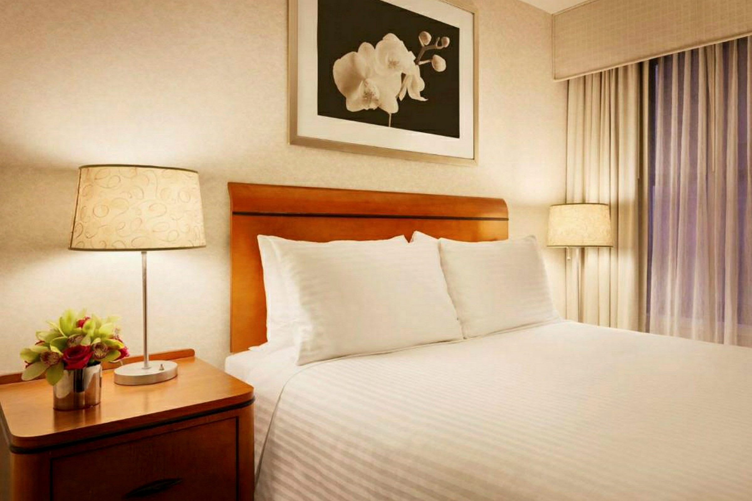 Et hotellrom med en stor seng, to puter, tredetaljer og et nattbord med en lampe og en vase med blomster. (AI-generert tekst)