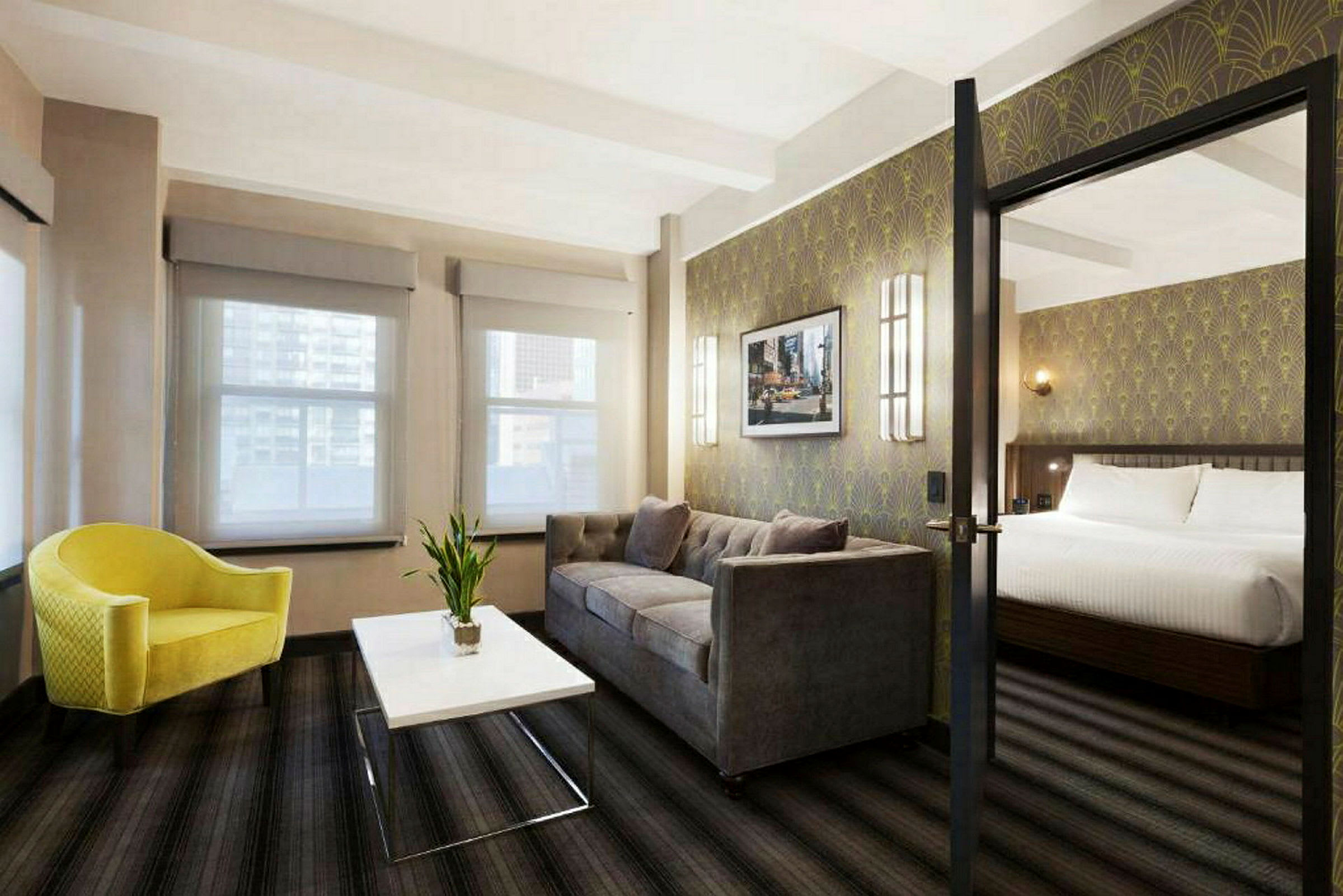 Et hotellrom med en sittegruppe bestående av en gul lenestol, et hvitt salongbord og en grå sofa. I bakgrunnen vises en soveplass med en dobbeltseng. (AI-generert tekst)