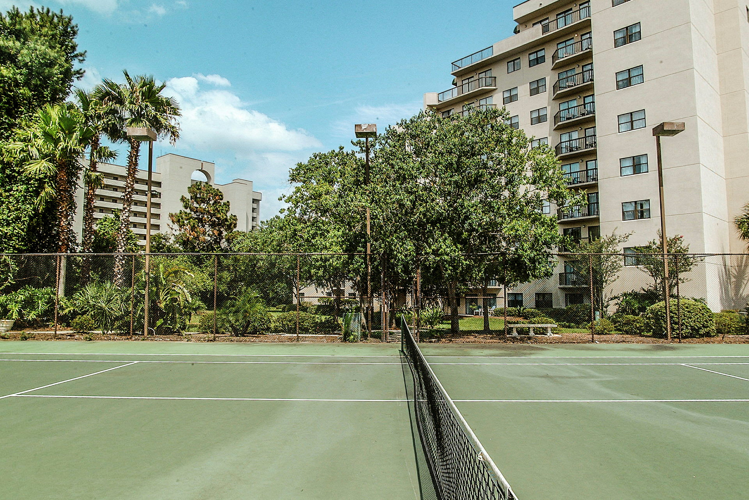 Tennisbane omgitt av grønne planter og bygninger i bakgrunnen. (AI-generert tekst)