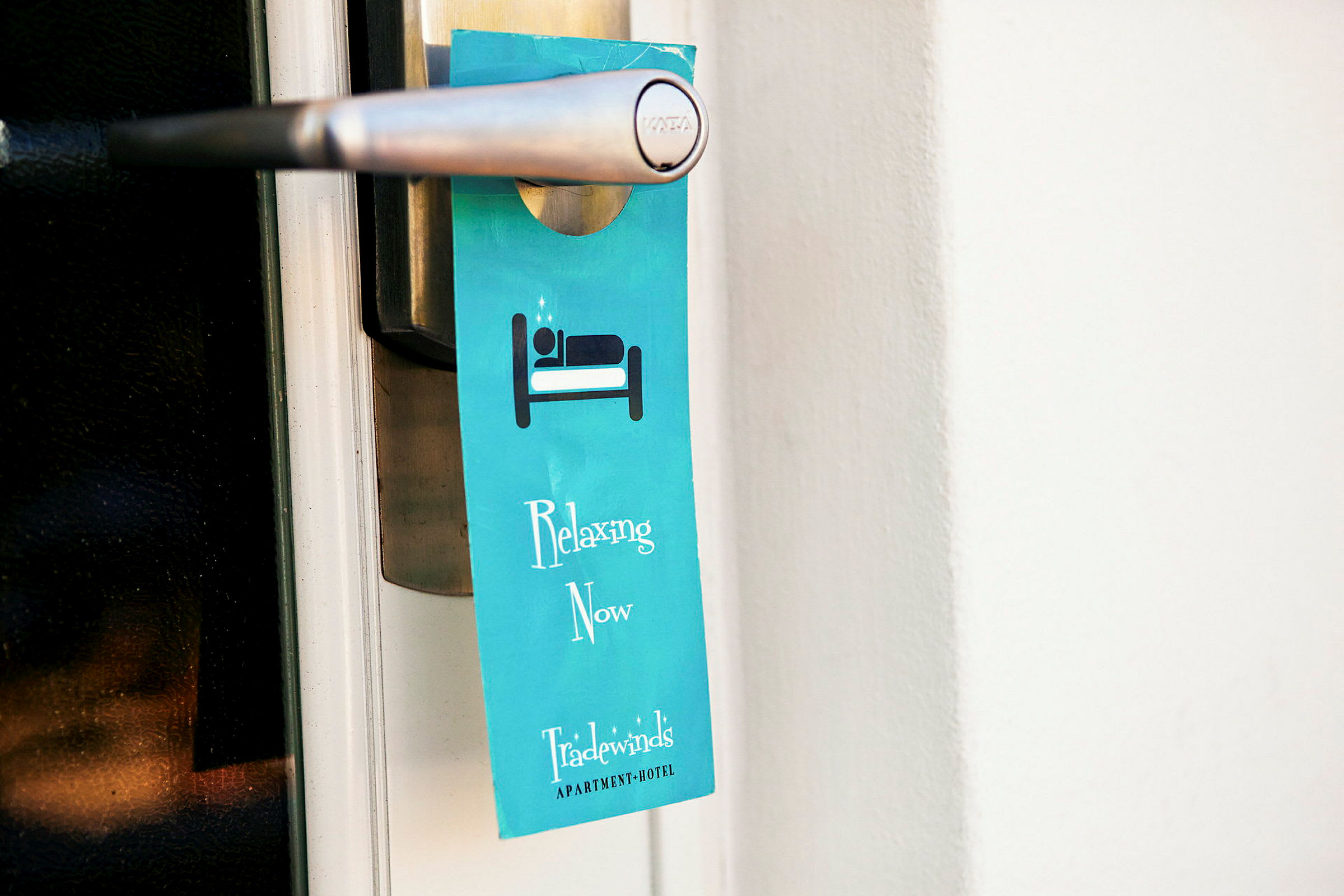 Et blått skilt henger på en dørhåndtak og viser teksten "Relaxing Now" samt hotellnavnet. (AI-generert tekst)