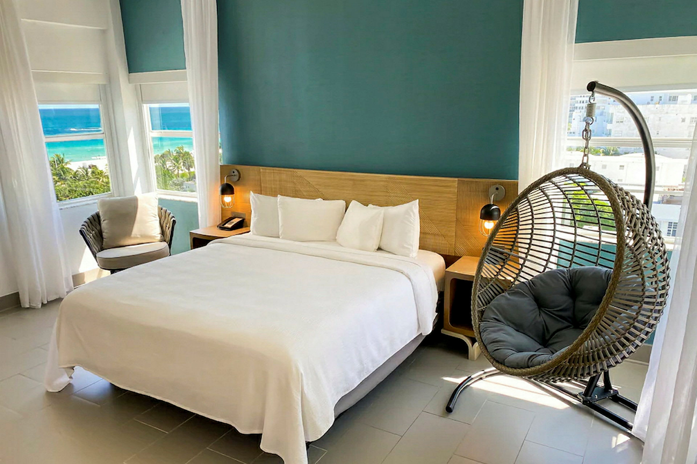 Et hotellrom med en dobbeltseng, en hengestol og store vinduer med utsikt over stranden. (AI-generert tekst)
