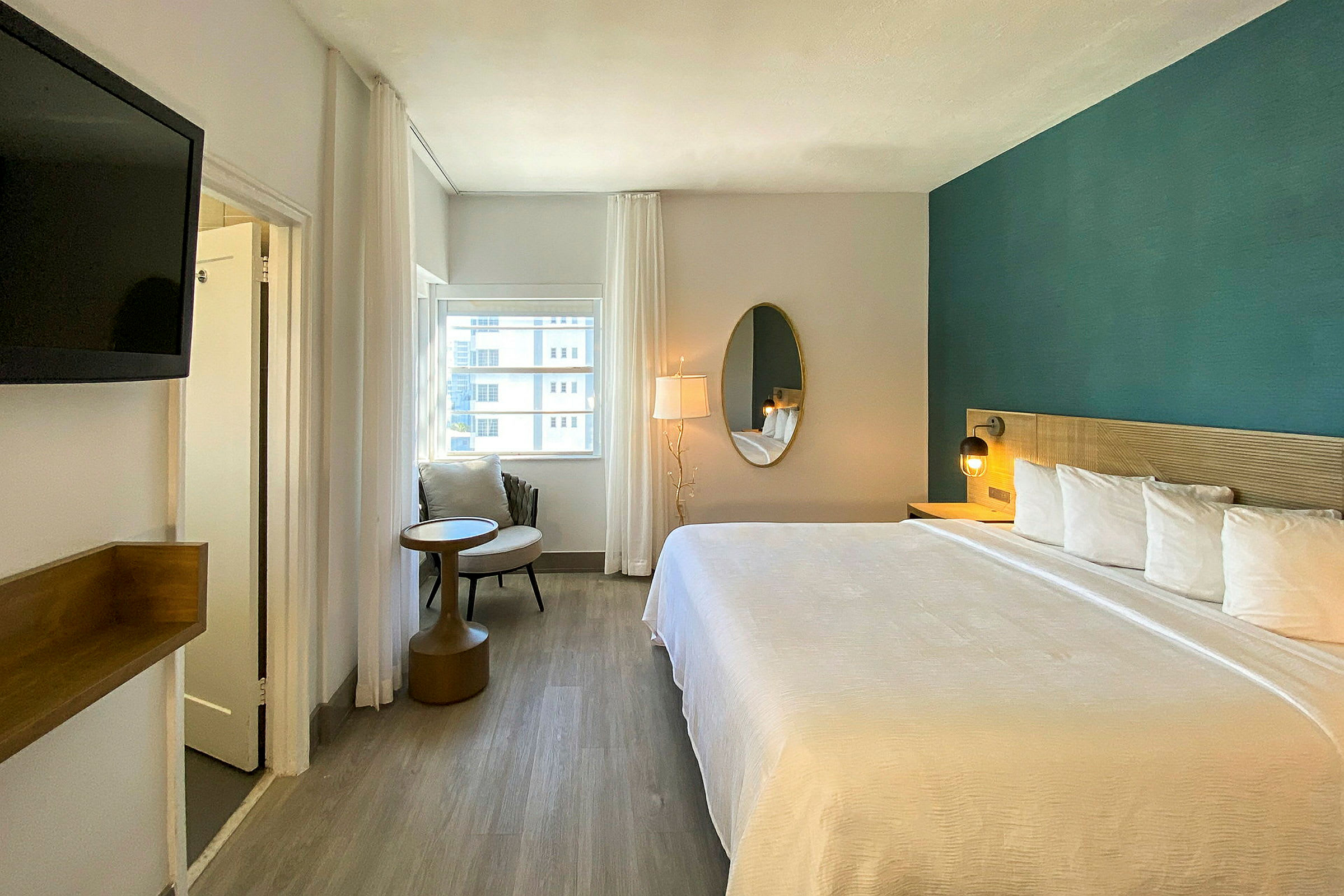 Moderne innredet hotellrom med stor seng, speil, lenestol og utsikt fra vinduet. (AI-generert tekst)