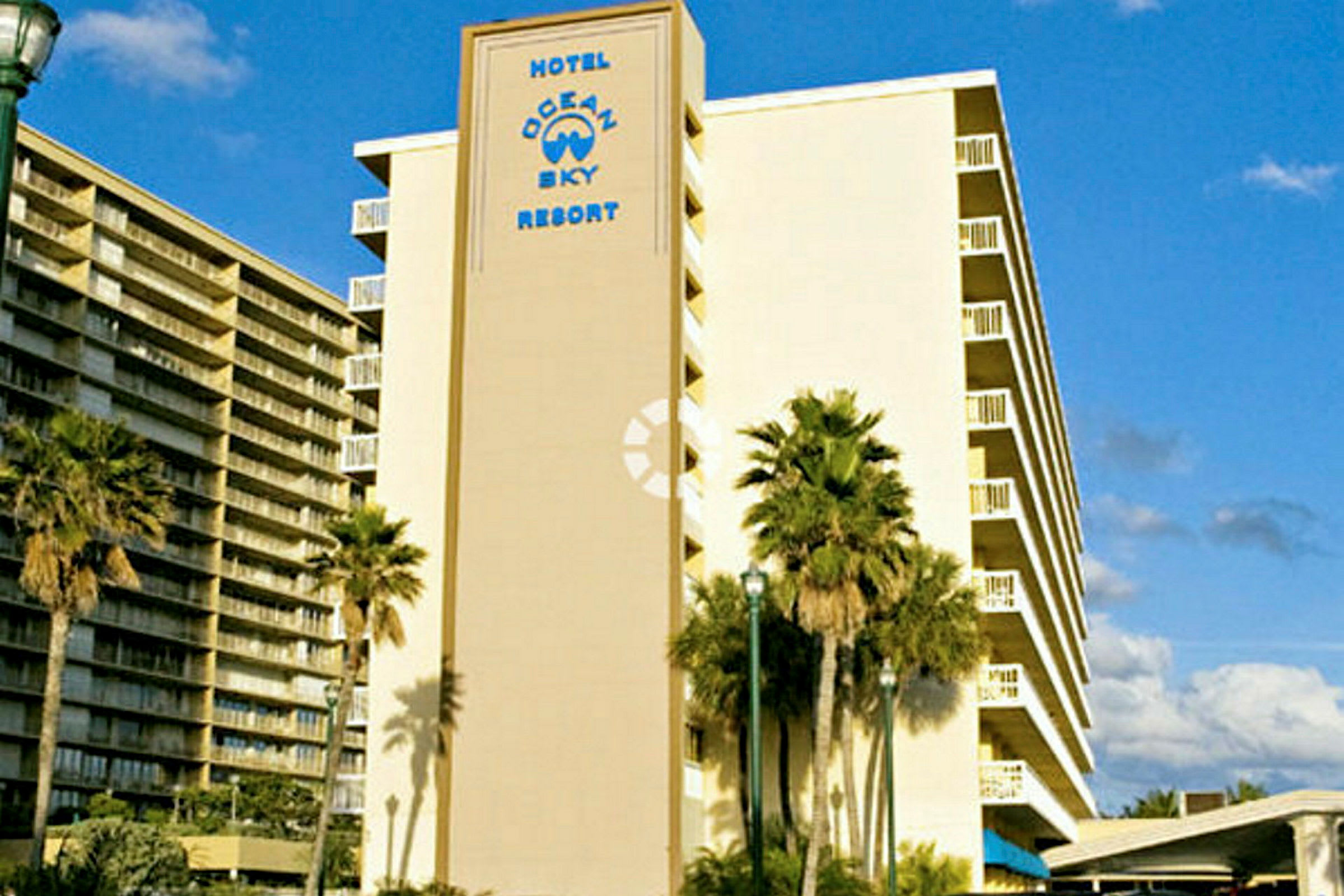 Et bilde av et hotell med teksten "Hotel Ocean Sky Resort" på fasaden. Palmer ses foran bygningen. (AI-generert tekst)