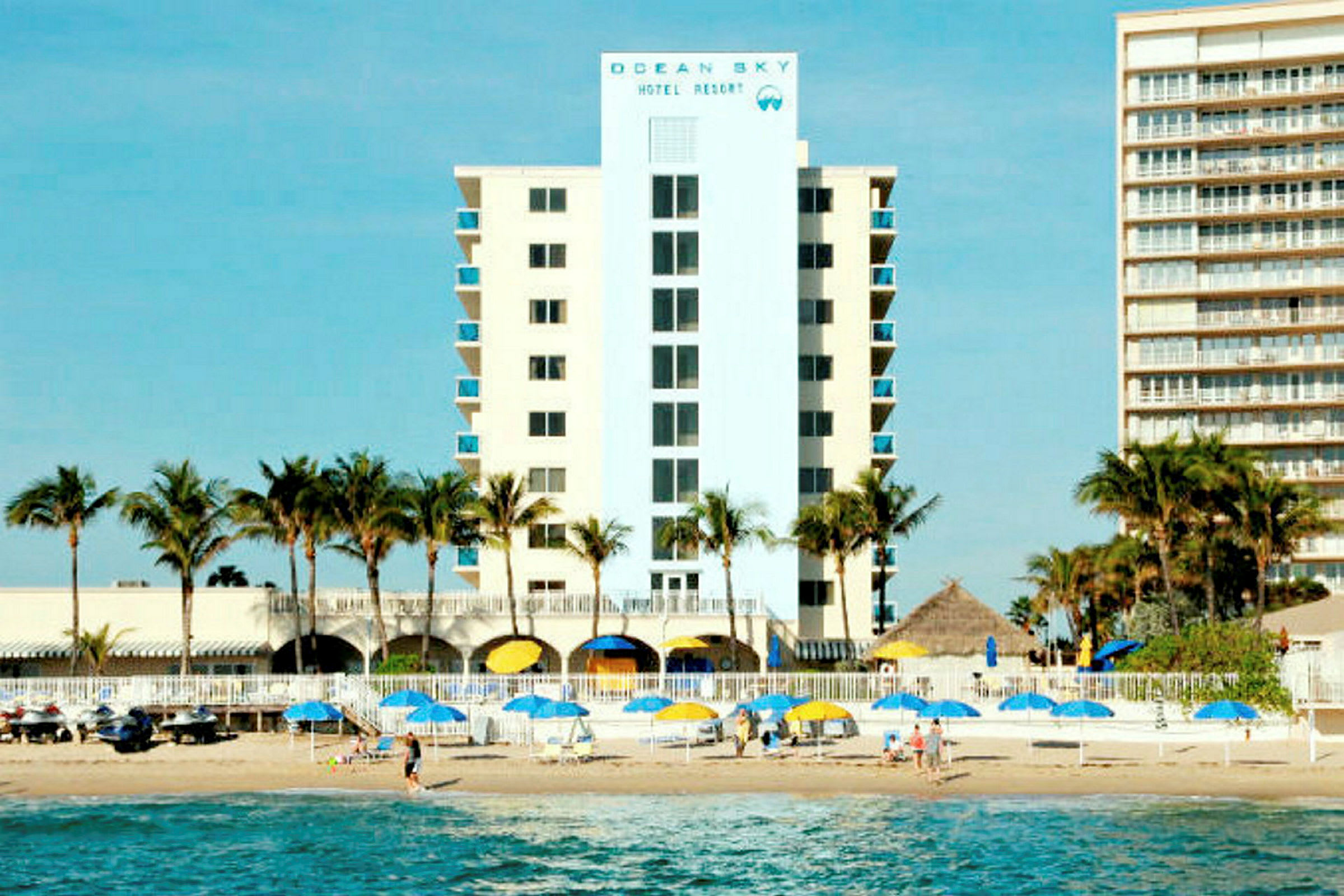 Et hotell med palmer foran, ved siden av stranden med parasoller og mennesker på sanden. (AI-generert tekst)