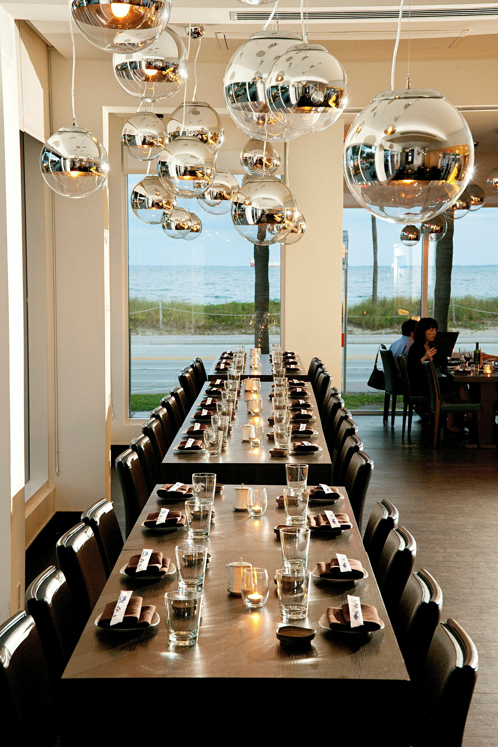 Bildet viser en restaurant med langbord som er dekket med glass og servietter. Taket har moderne metalliske lysekroner, og det er høye vinduer med utsikt over havet. (AI-generert tekst)