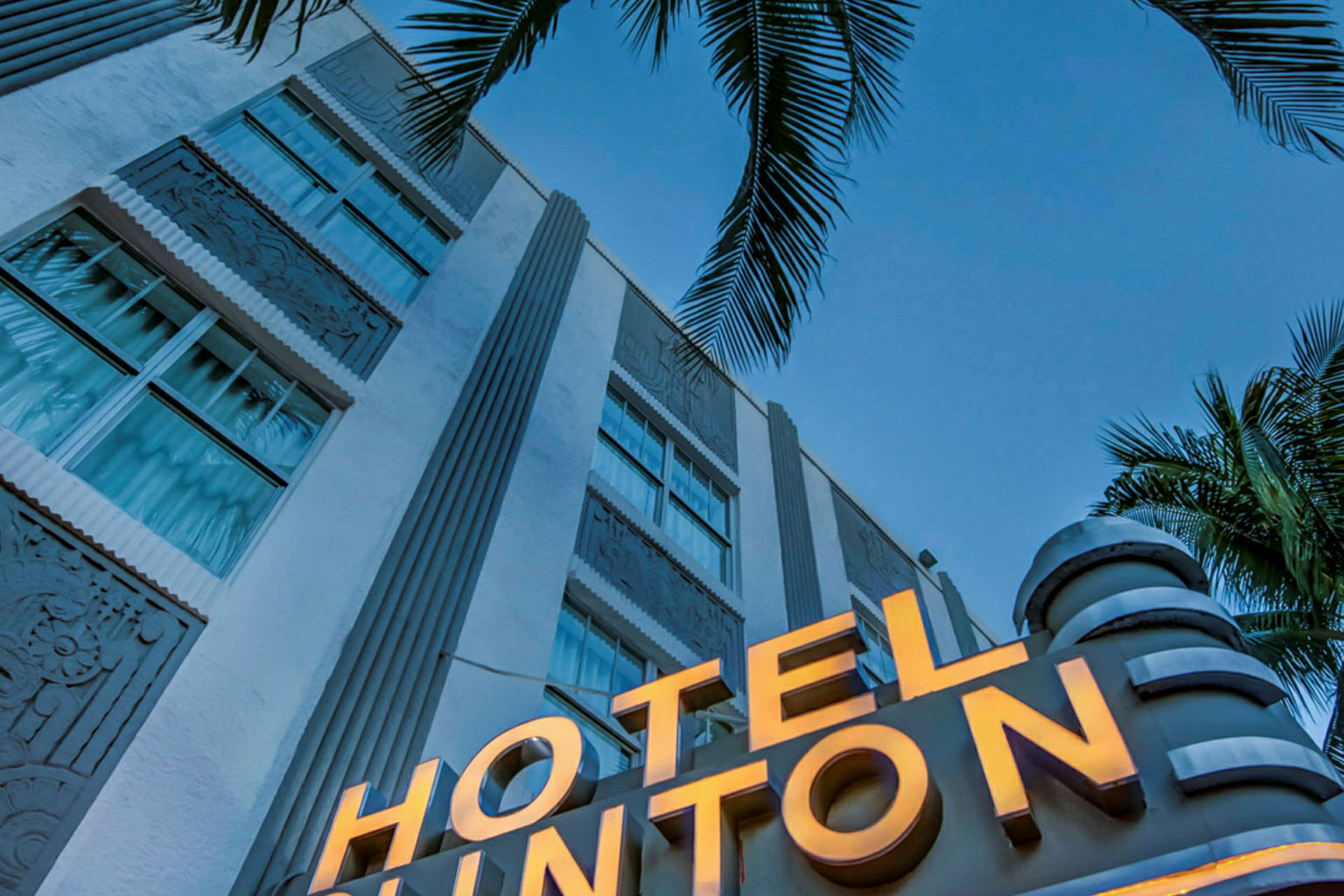 Et nærbilde av en hvit bygning med et opplyst skilt som sier "HOTEL" og palmer i forgrunnen mot en klar blå himmel. (AI-generert tekst)