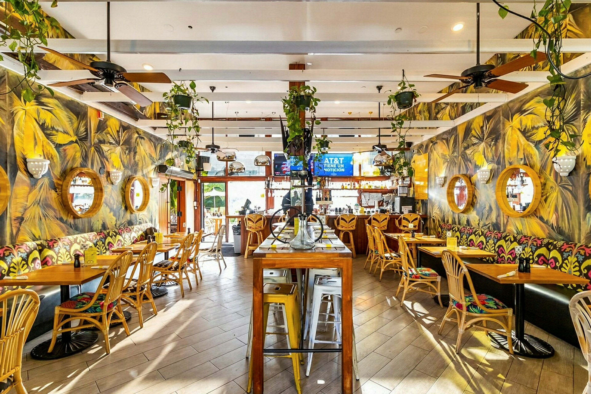 Interiøret i en restaurant med trestoler, bord og fargerike veggmalerier. Veggene er dekorert med runde speil, og planter henger fra taket. (AI-generert tekst)