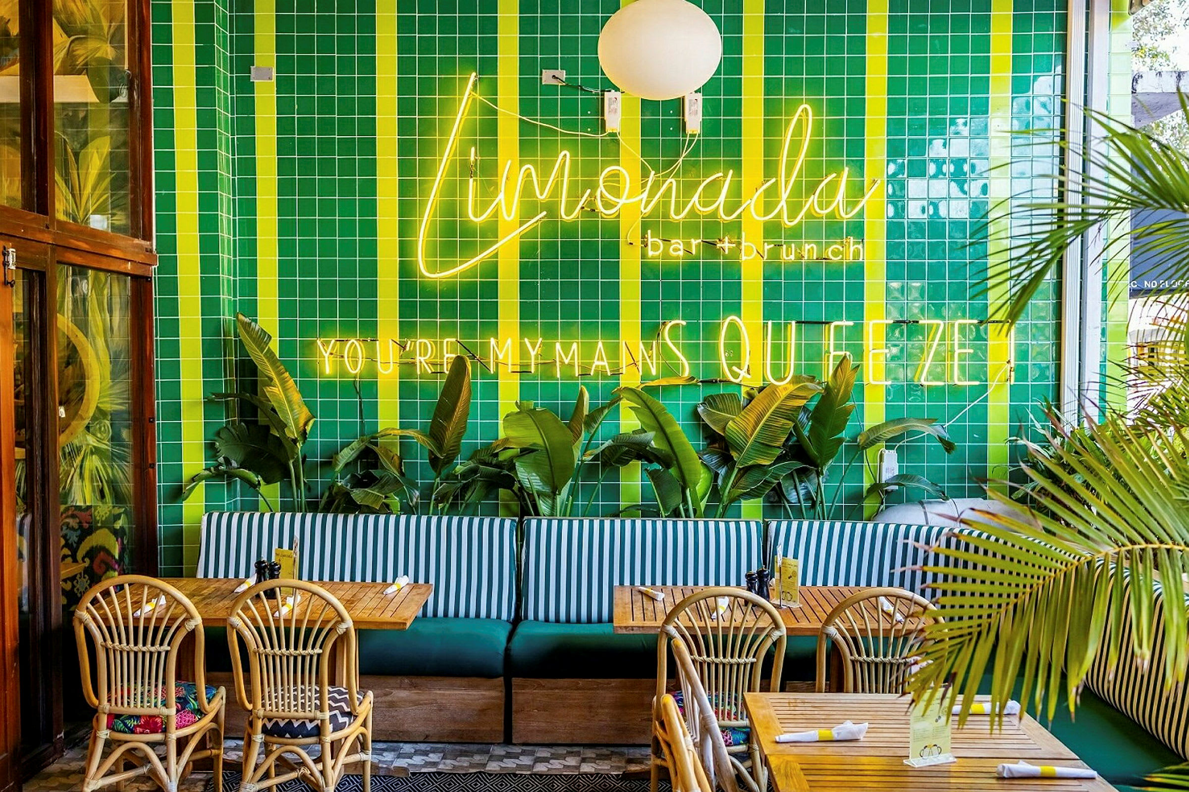 Restaurantinteriør med grønne flislagte vegger, neonskilt, palmer, trebord og flettede stoler. (AI-generert tekst)
