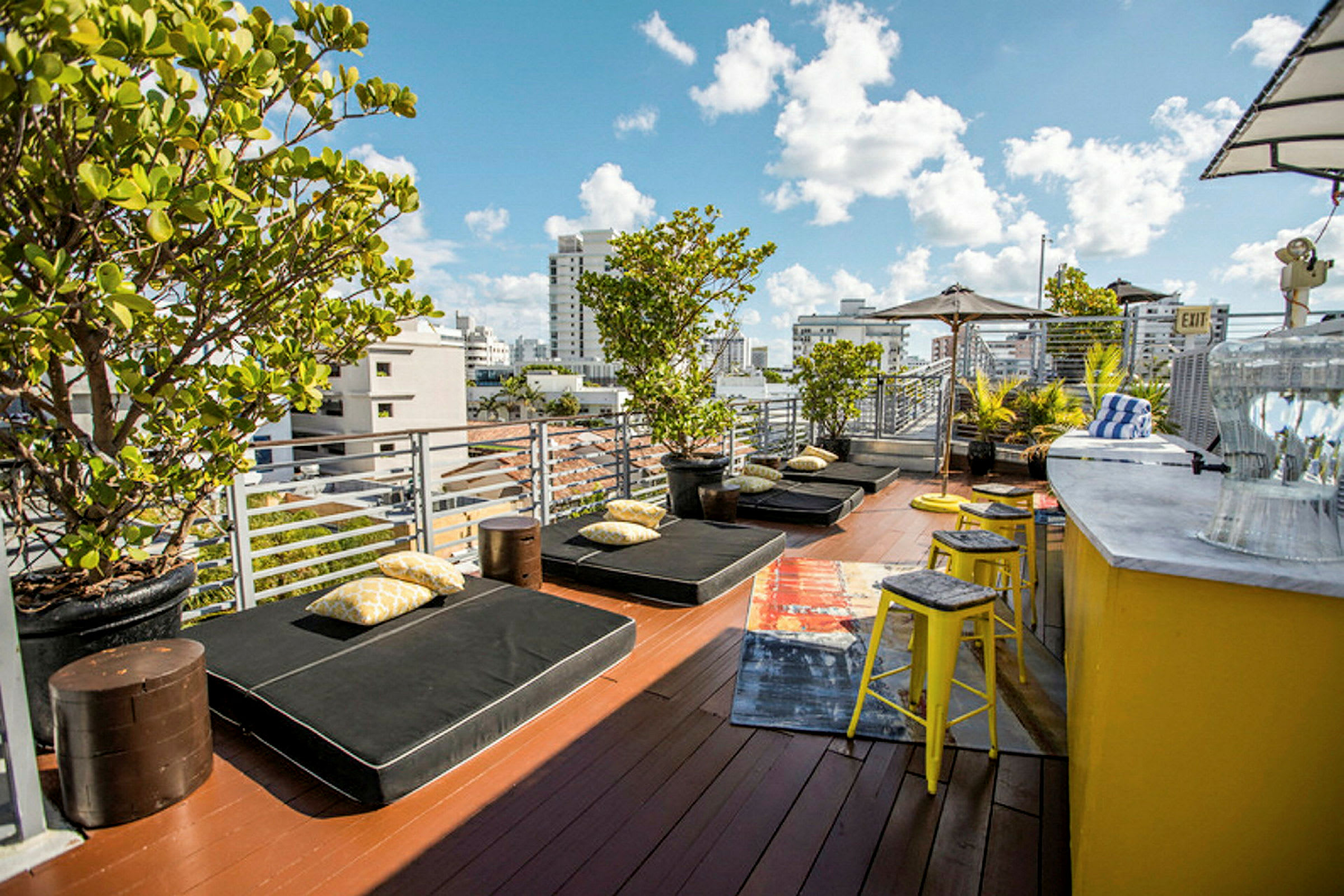 En terrasse med solsenger, puter og grønne planter. Det er en gul bardisk med krakker på siden. (AI-generert tekst)
