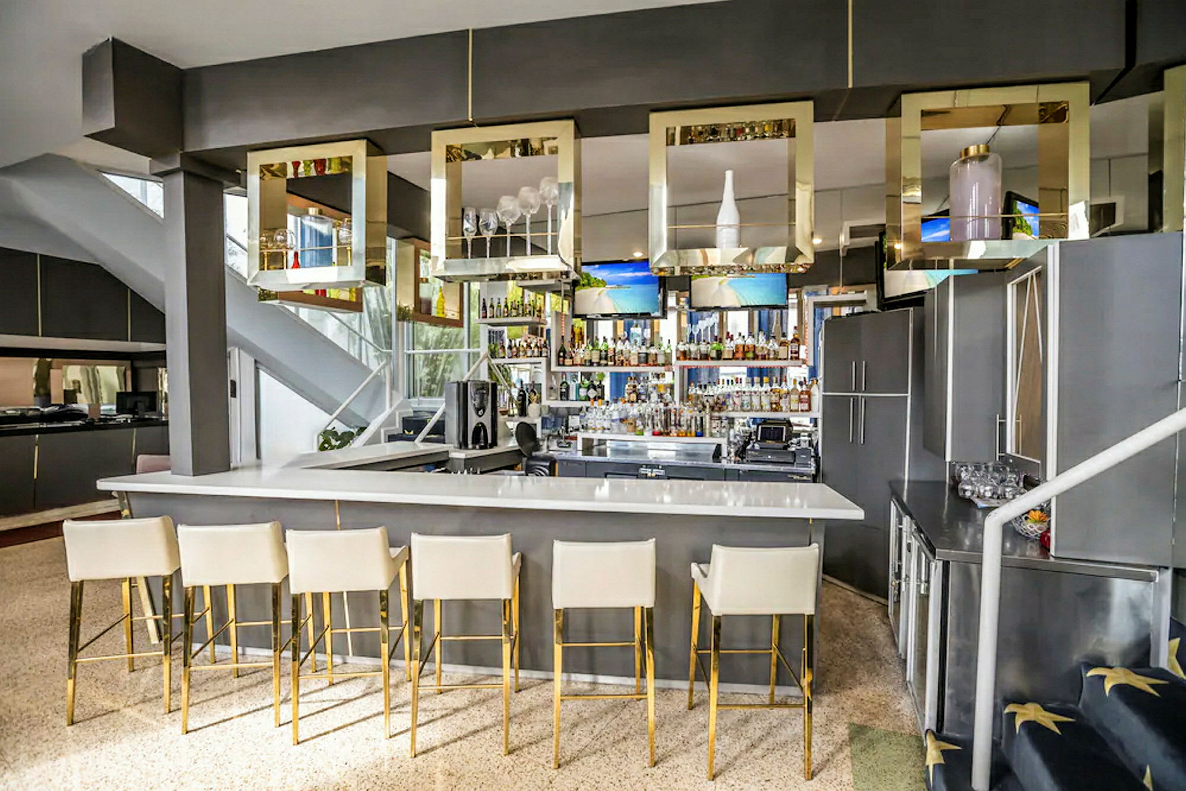 En moderne bar med hvit bardisk og høye stoler i lys farge med gyldne ben. Over bardisken henger firkantede dekorasjoner. (AI-generert tekst)