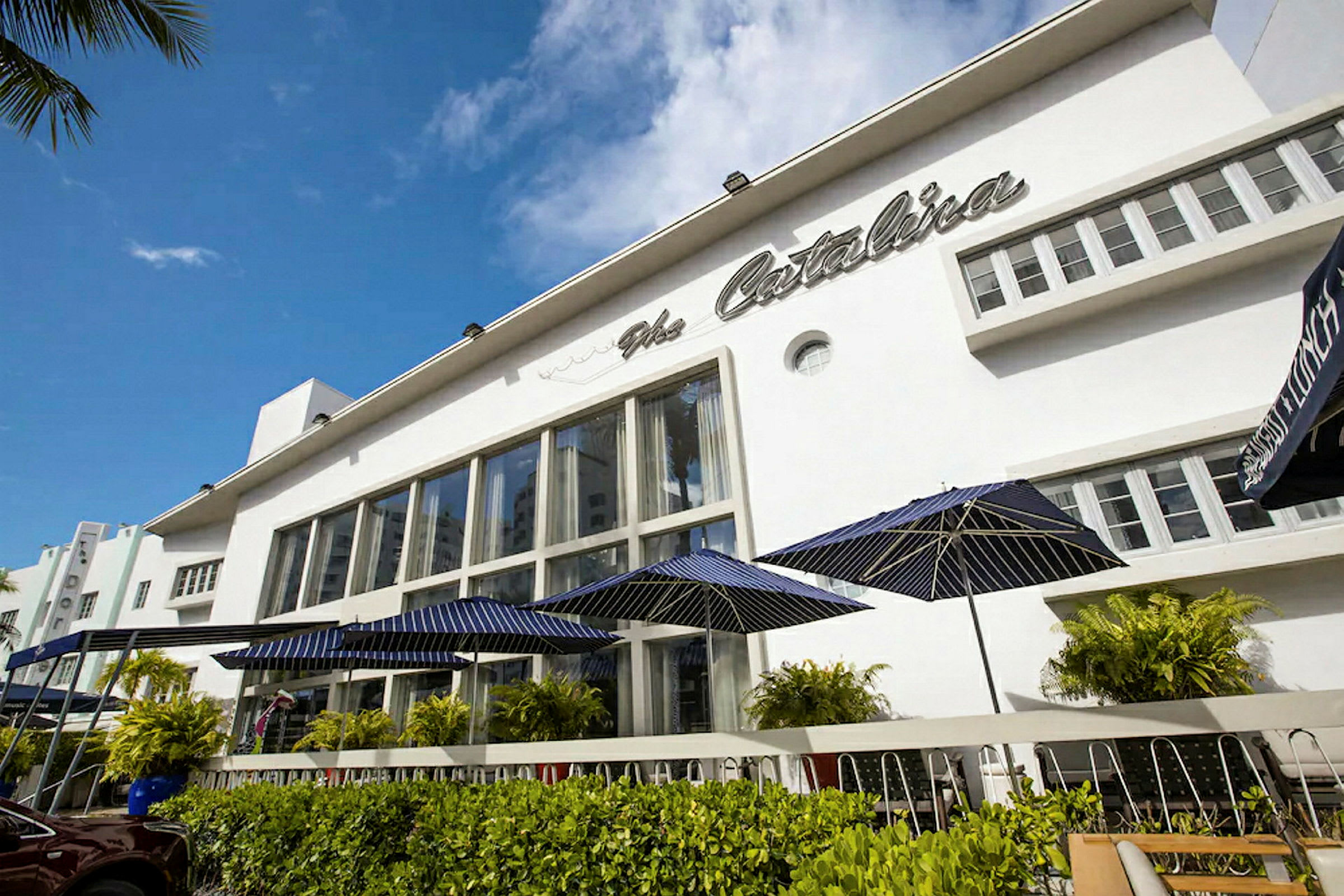 En bygning med store vinduer og navnet "The Catalina" på fasaden. Foran bygningen er det grønne busker og bord med blå parasoller. (AI-generert tekst)
