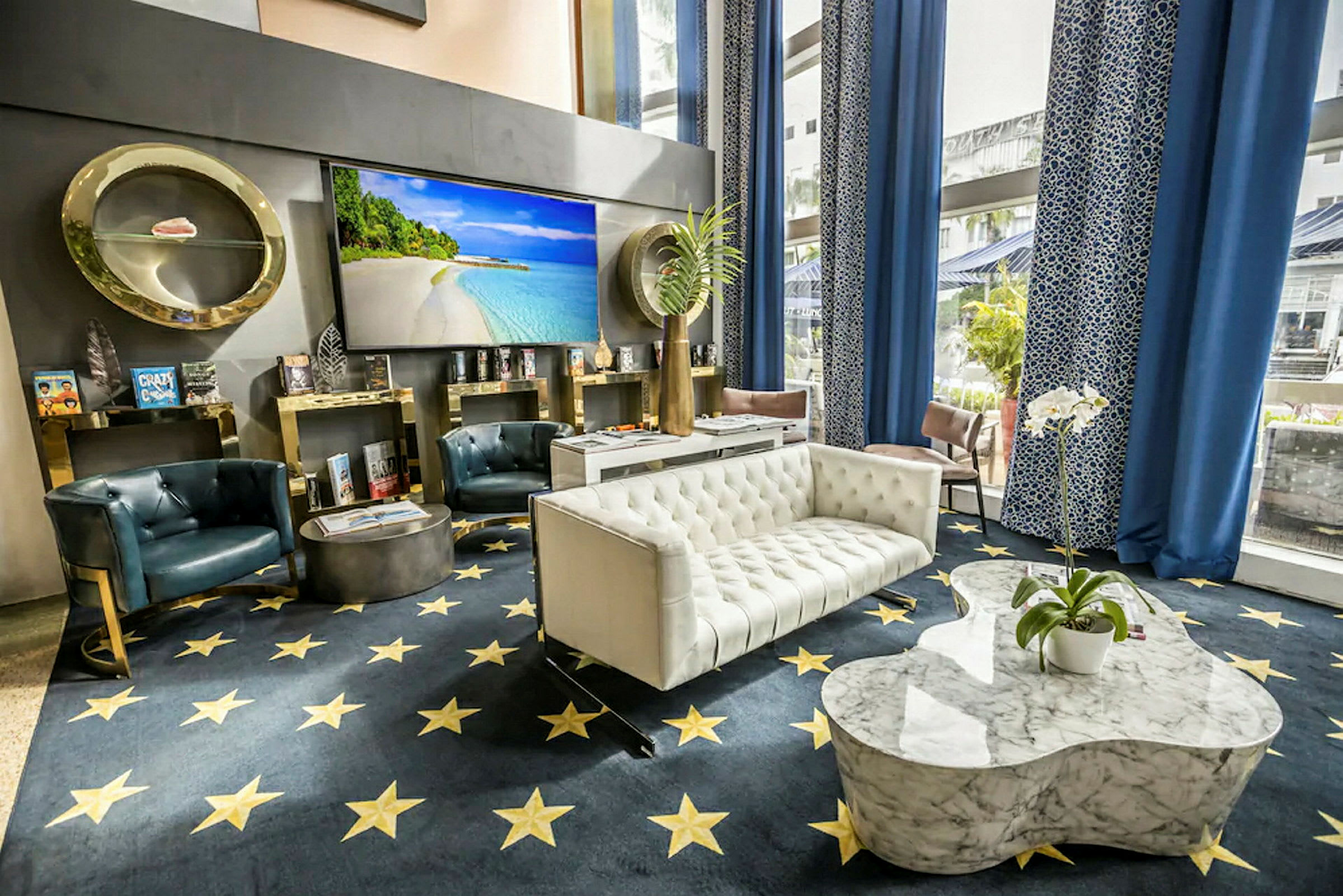 Bilde av en elegant hotellobby med sofaer, lenestoler og et marmorbord. Bakgrunnen har en stor skjerm med strandmotiv, og interiøret inkluderer blå gardiner og dekorative detaljer. (AI-generert tekst)