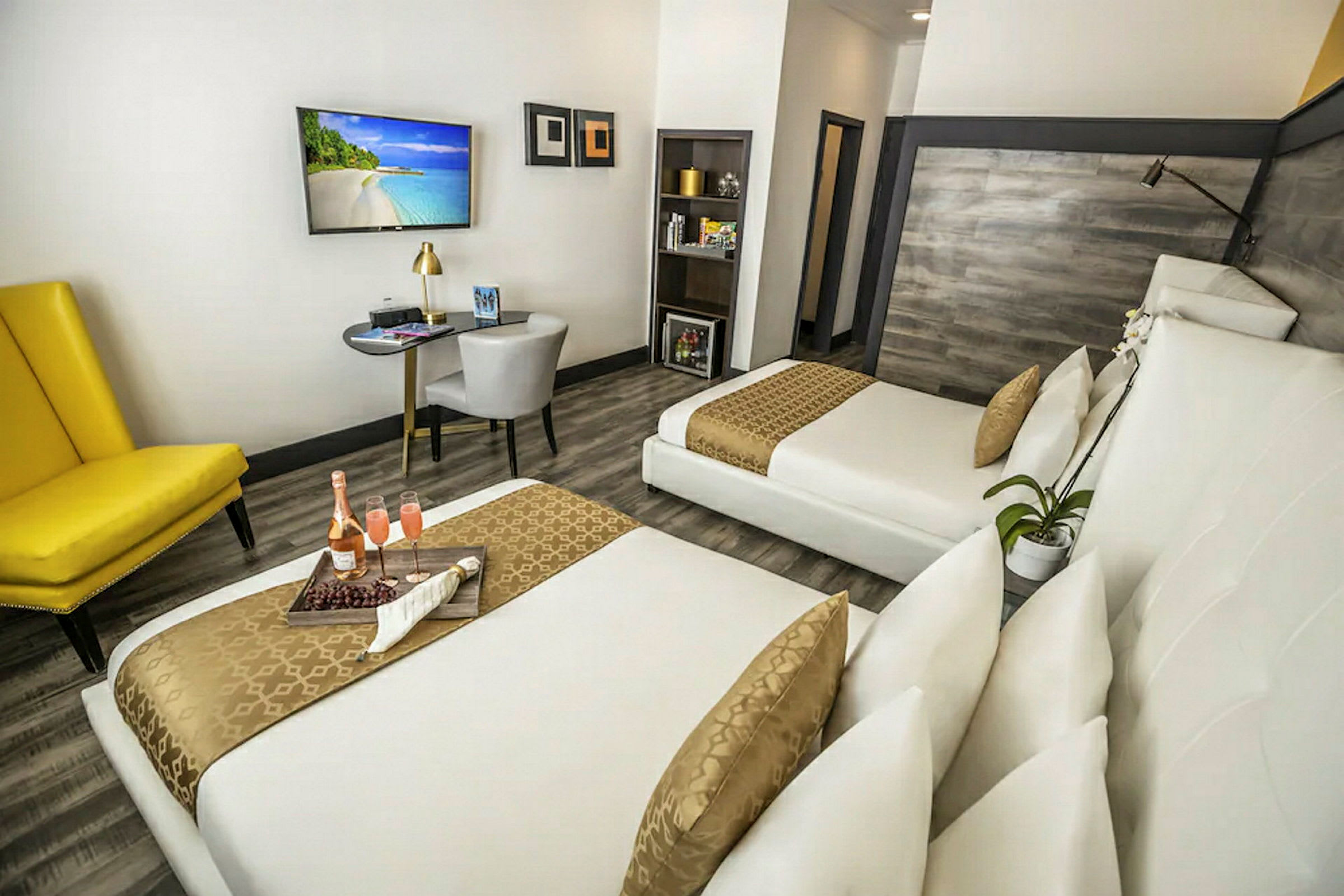 Et moderne hotellrom med to senger, en gul lenestol, et skrivebord og en flatskjerm-TV på veggen. (AI-generert tekst)