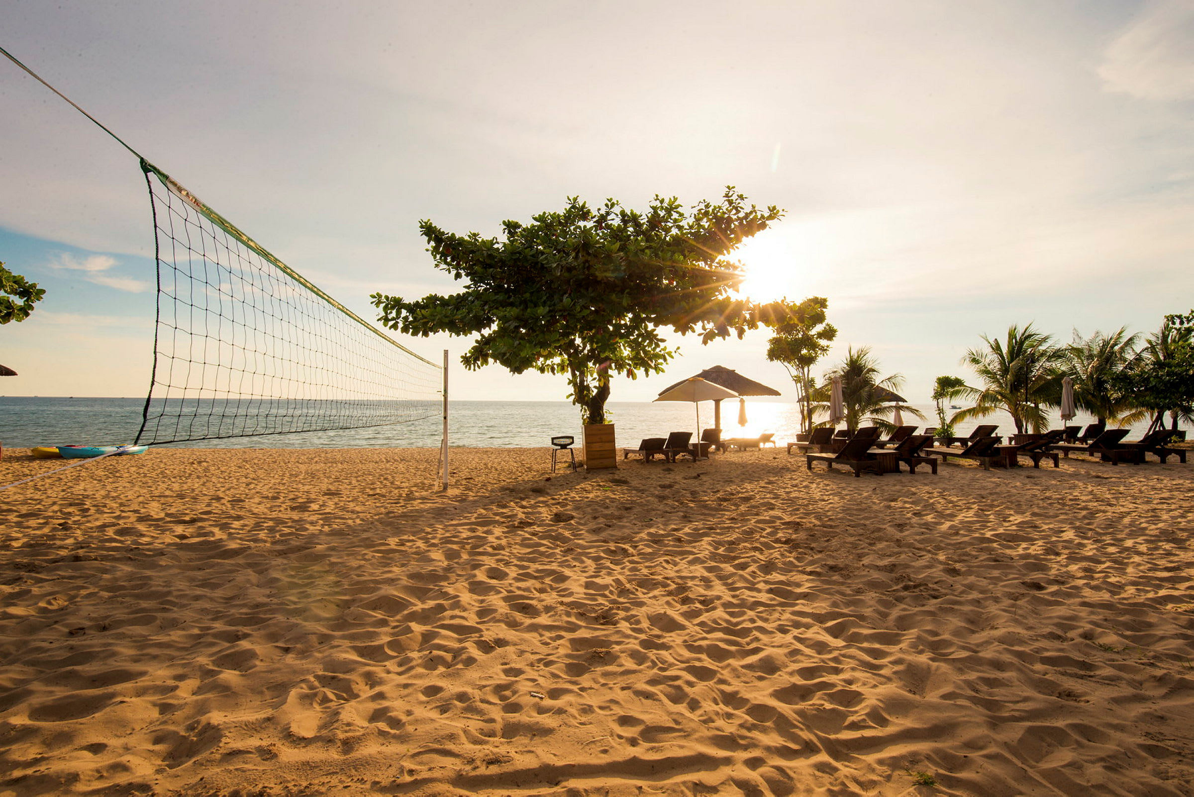 En sandstrand med et volleyballnett, solsenger under parasoller, og noen trær. Havet er synlig i bakgrunnen under en solnedgang. (AI-generert tekst)