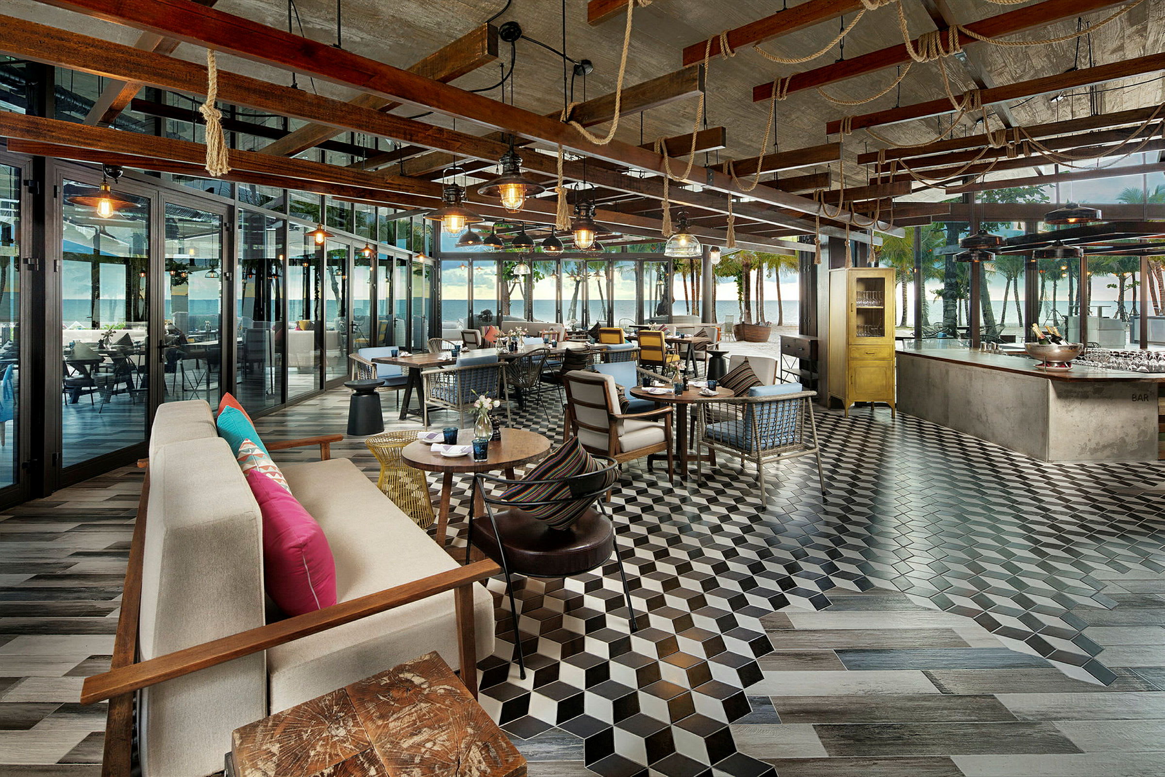 En kafé eller restaurant med moderne interiør, sittegrupper og bord, samt store glasspartier som vender ut mot en terrasse med utsikt over havet og palmer. (AI-generert tekst)