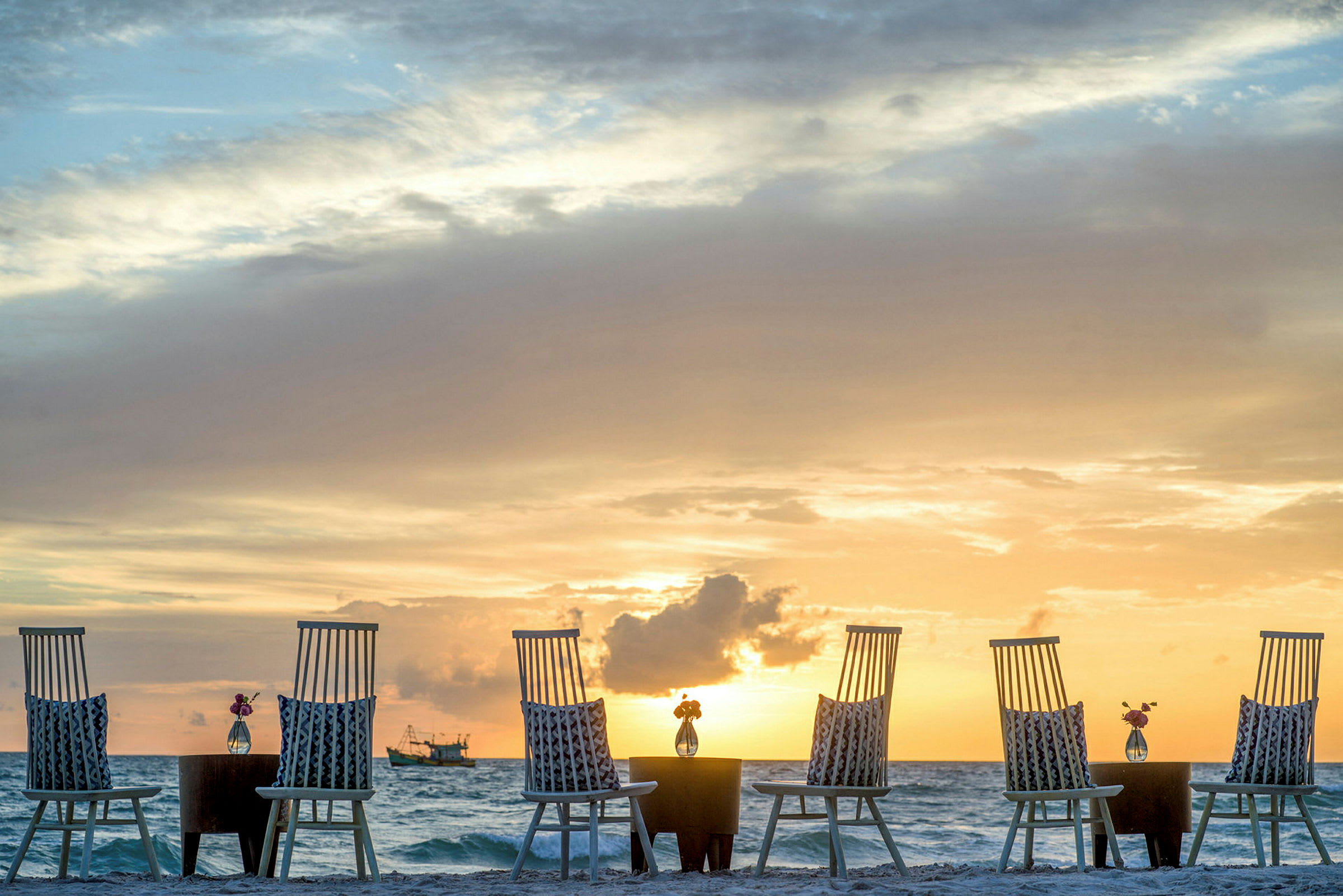 Stoler og bord på en strand med havet og solnedgang i bakgrunnen. (AI-generert tekst)