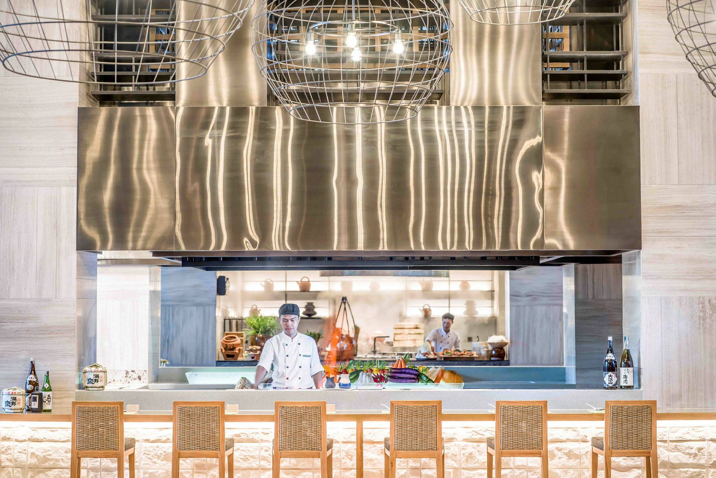 En moderne restaurant med åpent kjøkkenmiljø. En kokk jobber bak en stor, utstillingslignende benk hvor grønnsaker og flasker står fremme. En metallisk vifte ovenfor. (AI-generert tekst)