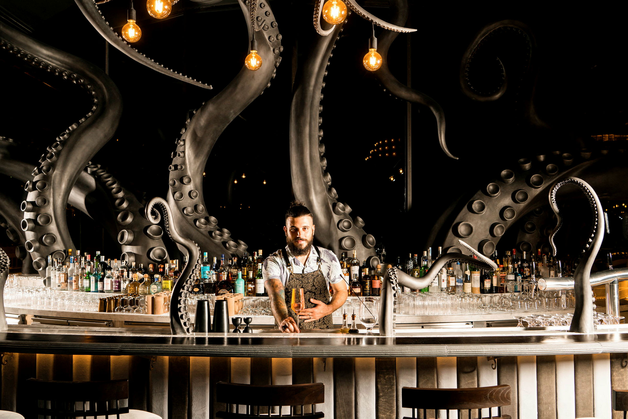 En bartender står ved en bar med detaljer av blekksprut-tentakler, flasker og glass vises bak ham. (AI-generert tekst)