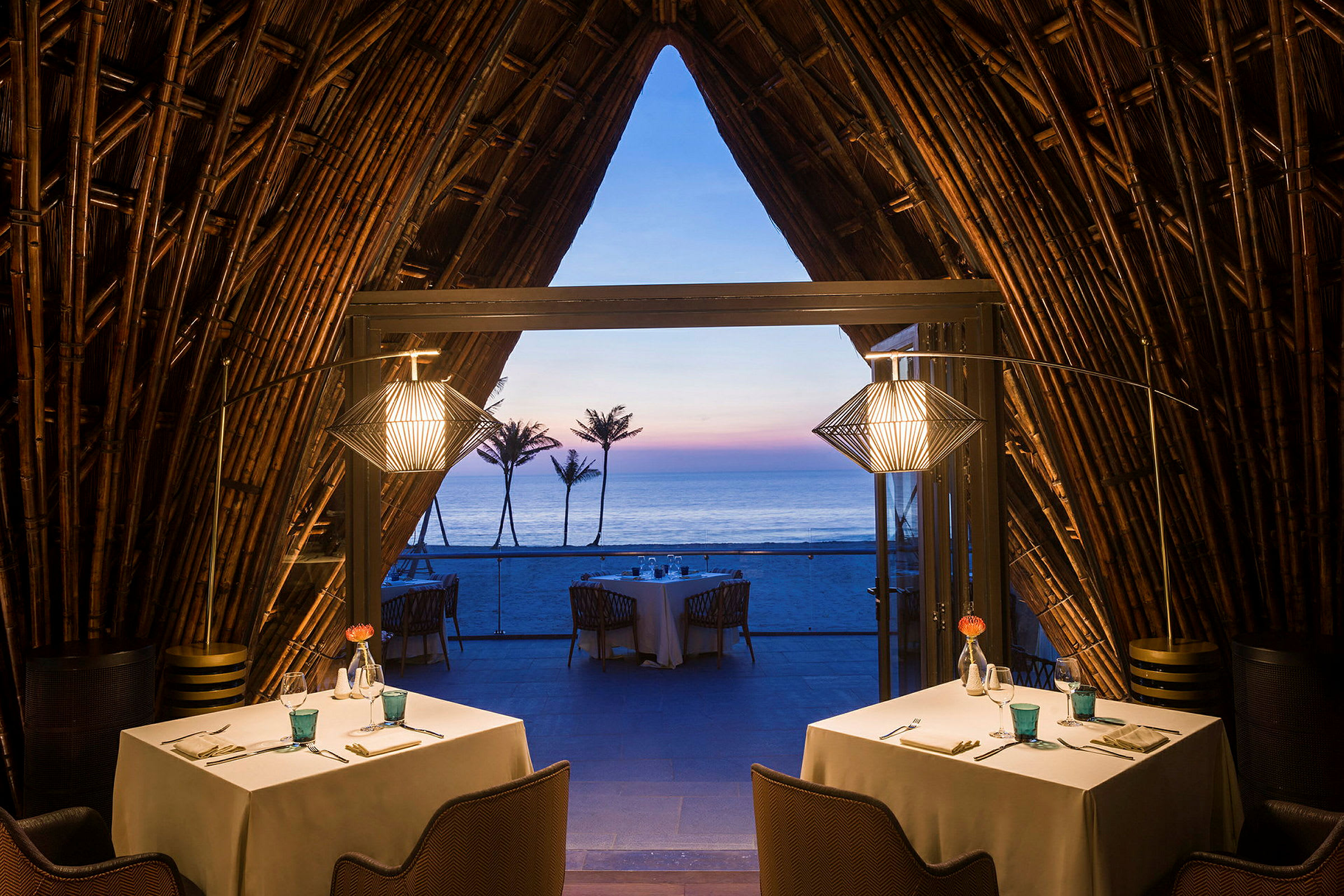 Interiør av en restaurant med borddekking, med åpen utsikt mot havet og solnedgangen. Trekonstruksjoner omgir miljøet. (AI-generert tekst)