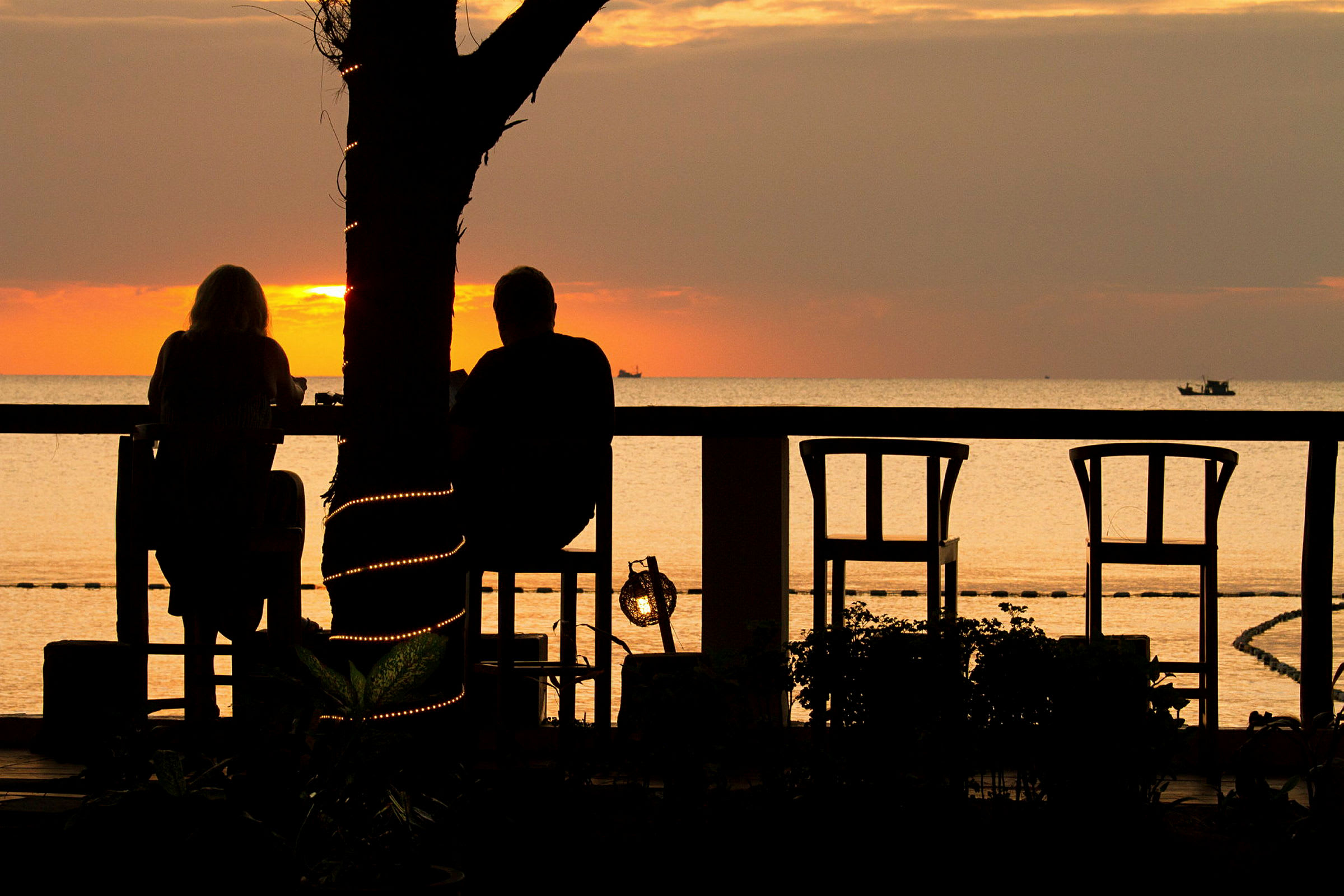 To personer sitter ved et bord og nyter solnedgangen ved havet. Et tre med dekorative lys står i forgrunnen. (AI-generert tekst)