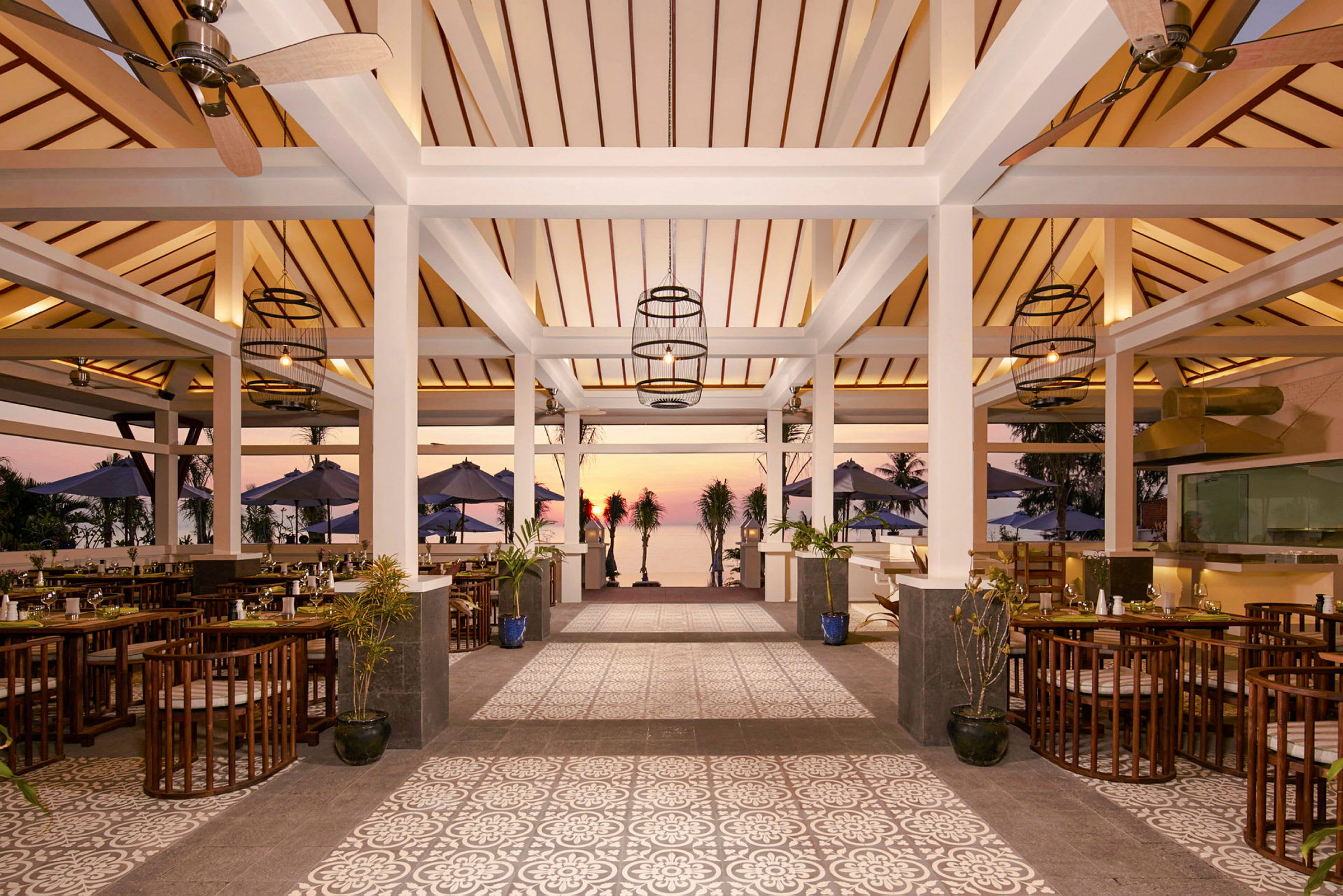En restaurant med tretak, åpne vegger og gulv med dekorative mønstre. Utenfor vises bord, stoler og parasoller samt solnedgang ved sjøen. (AI-generert tekst)