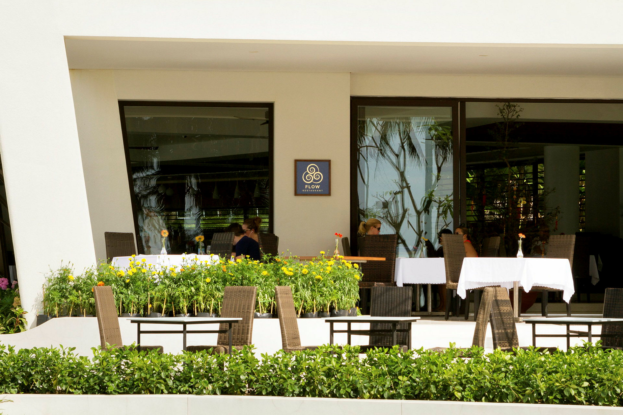 En restaurant med bord og stoler på en terrasse omgitt av grønne planter og blomster. (AI-generert tekst)