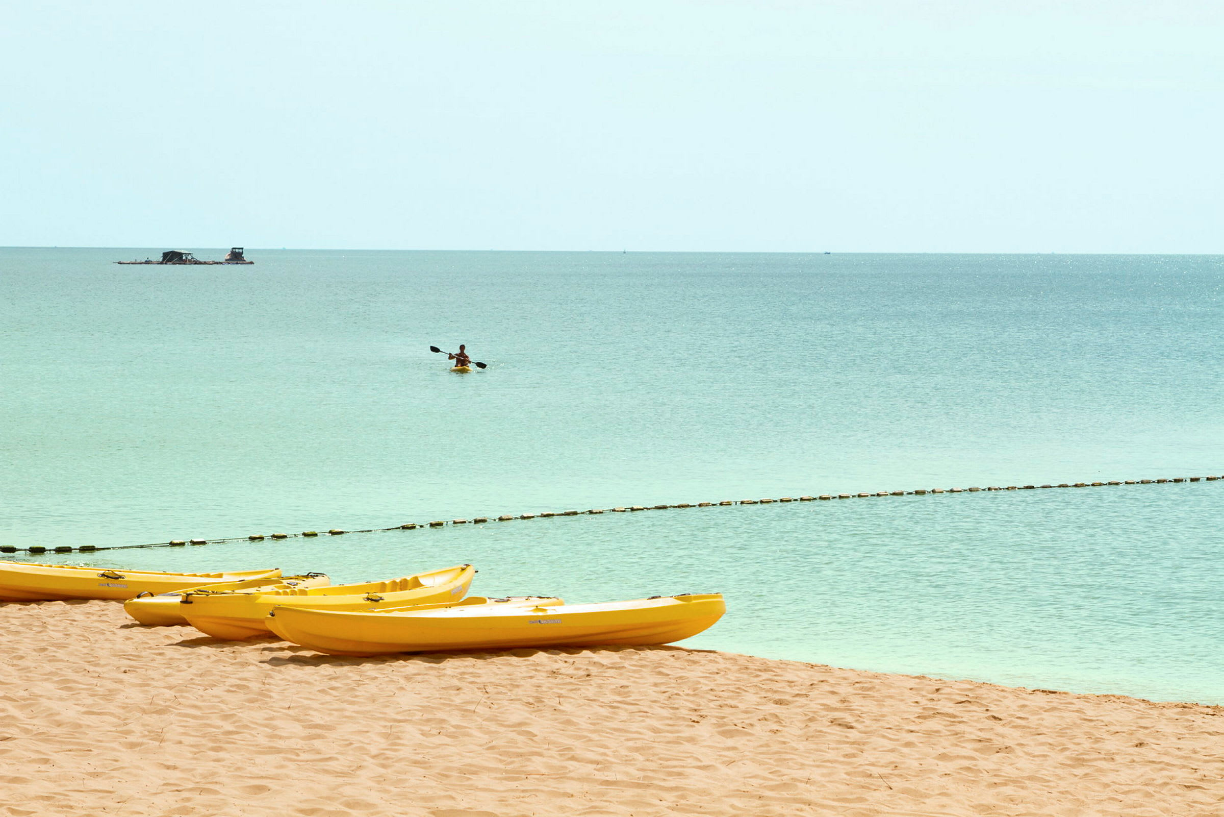 Flere gule kajakker ligger på en sandstrand, mens en person padler en kajakk på det rolige havet. (AI-generert tekst)