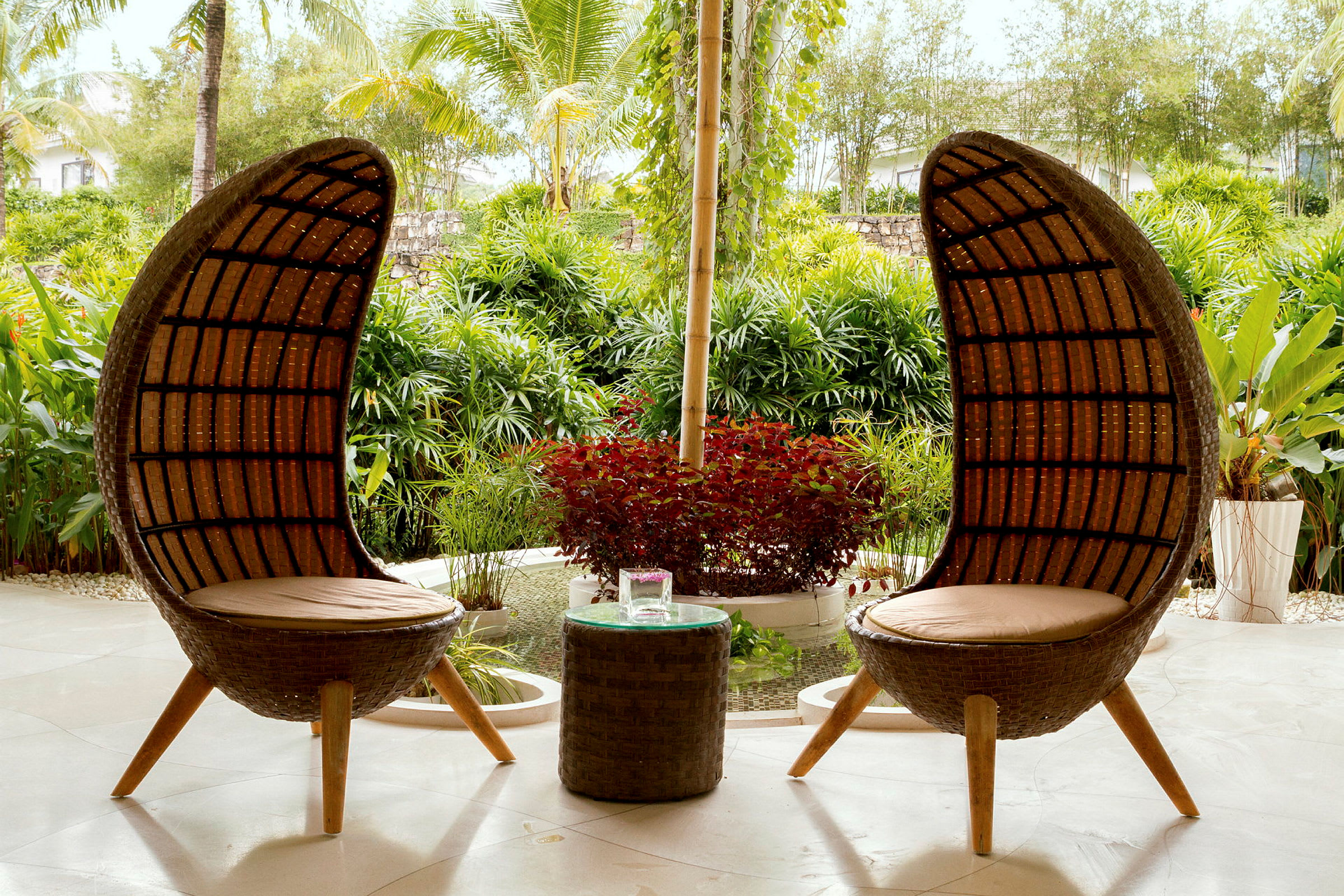 To rottingstoler med høye ryggstøtter og et lite bord står på en terrasse omgitt av frodig grønt. (AI-generert tekst)