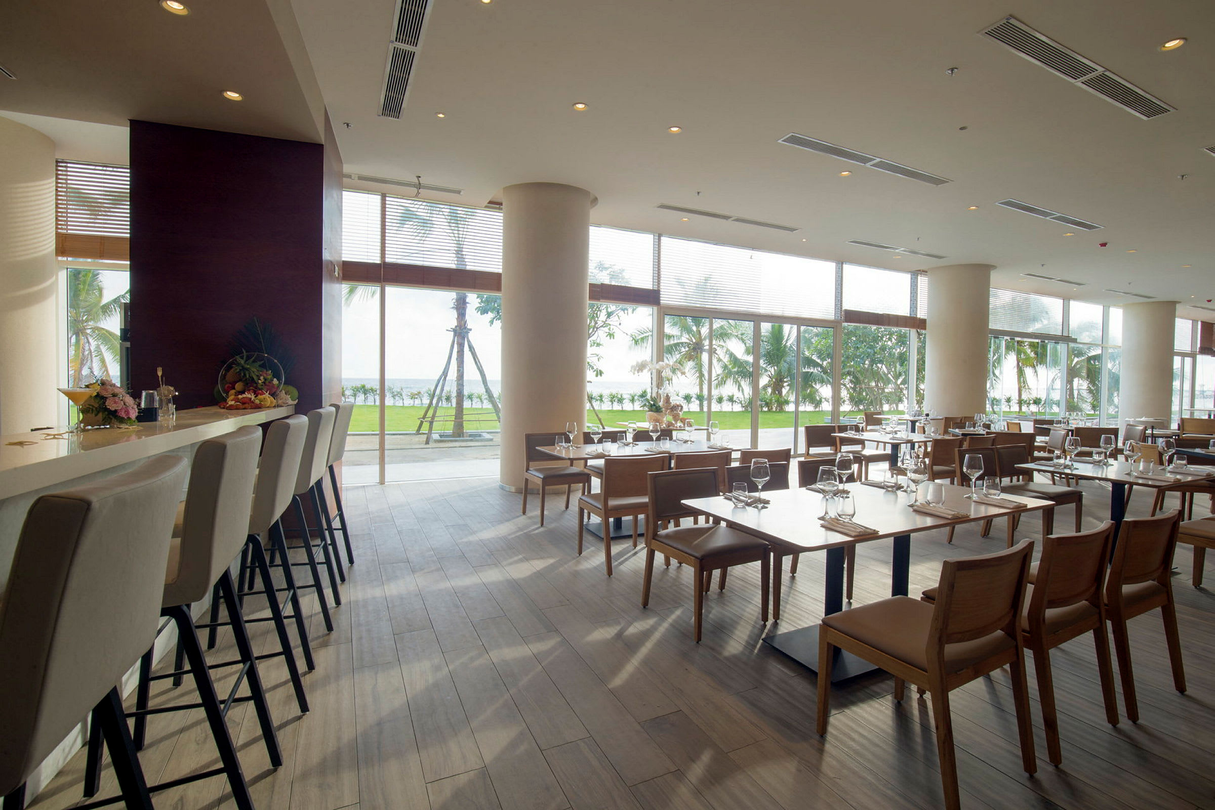 Interiøret av en lys restaurant med bord og stoler, store vinduer med utsikt mot grønne områder og palmer utenfor. (AI-generert tekst)