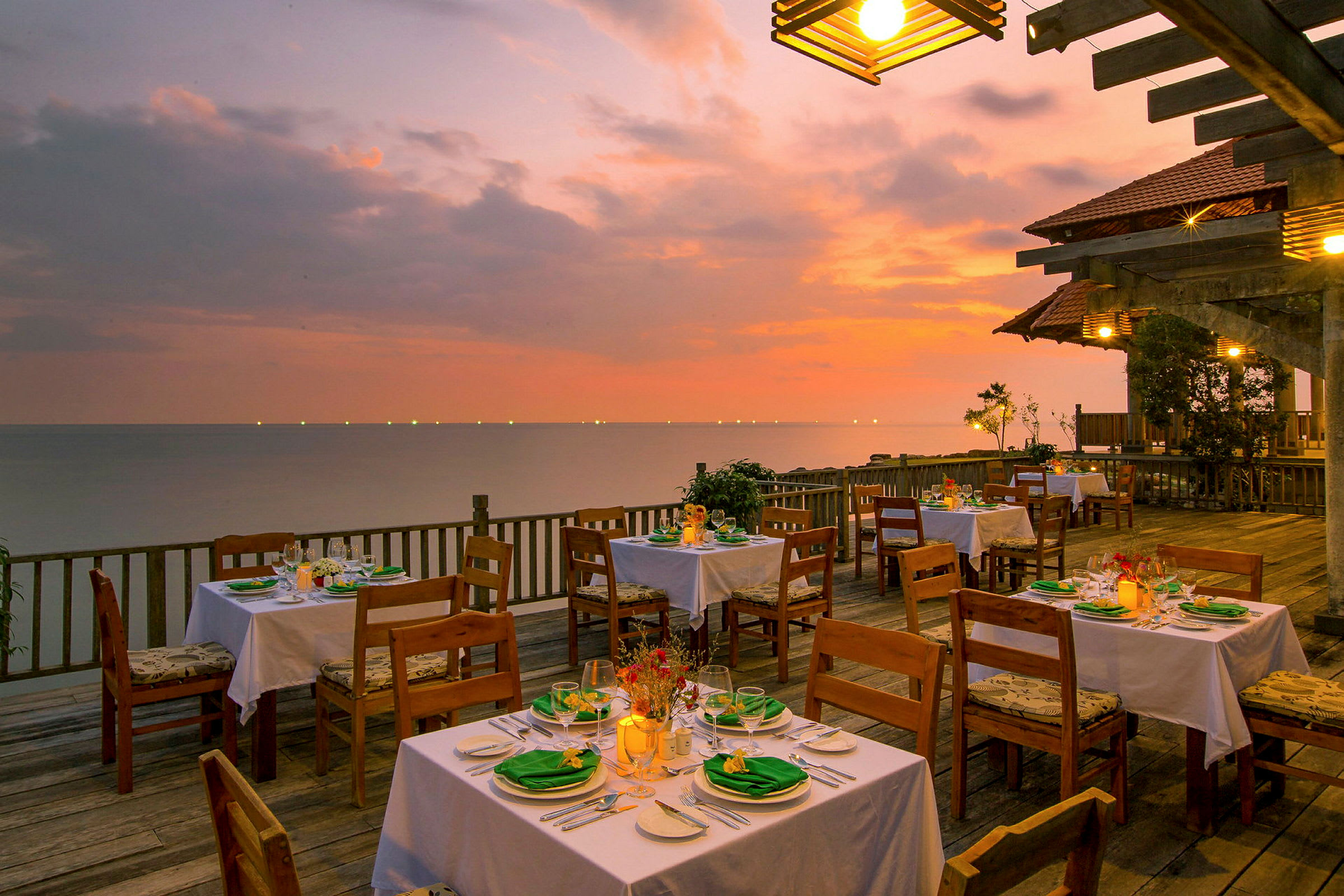 En uteservering med dekkede bord på en treterrasse ved havet under en fargerik solnedgang. (AI-generert tekst)
