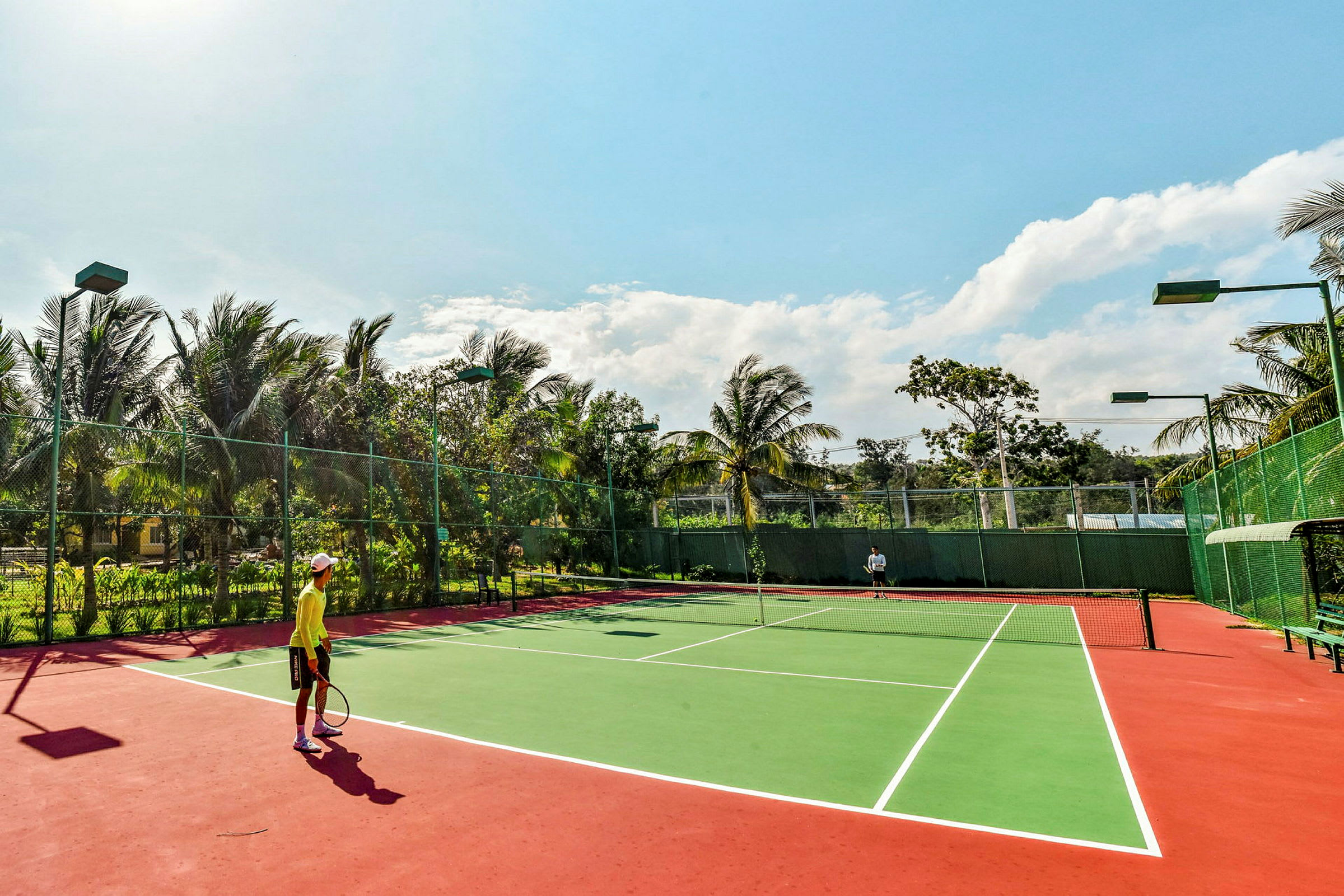 To personer spiller tennis på en utendørs bane omgitt av vegetasjon og palmer under en klar himmel. (AI-generert tekst)
