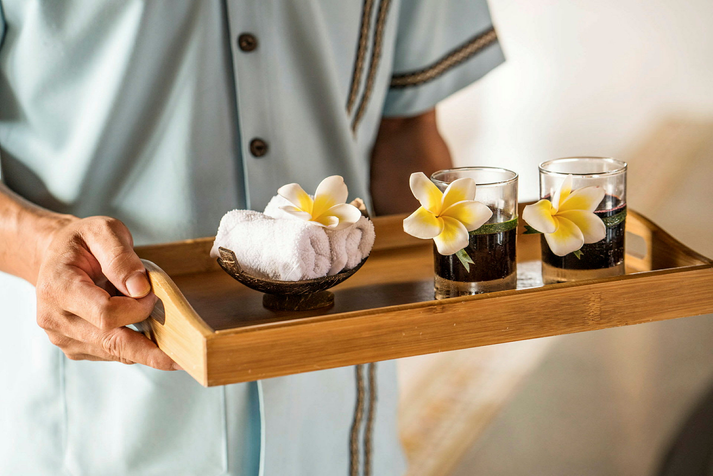 En person holder et trebord med to glass dekorert med blomster og en skål med rullede håndklær. (AI-generert tekst)