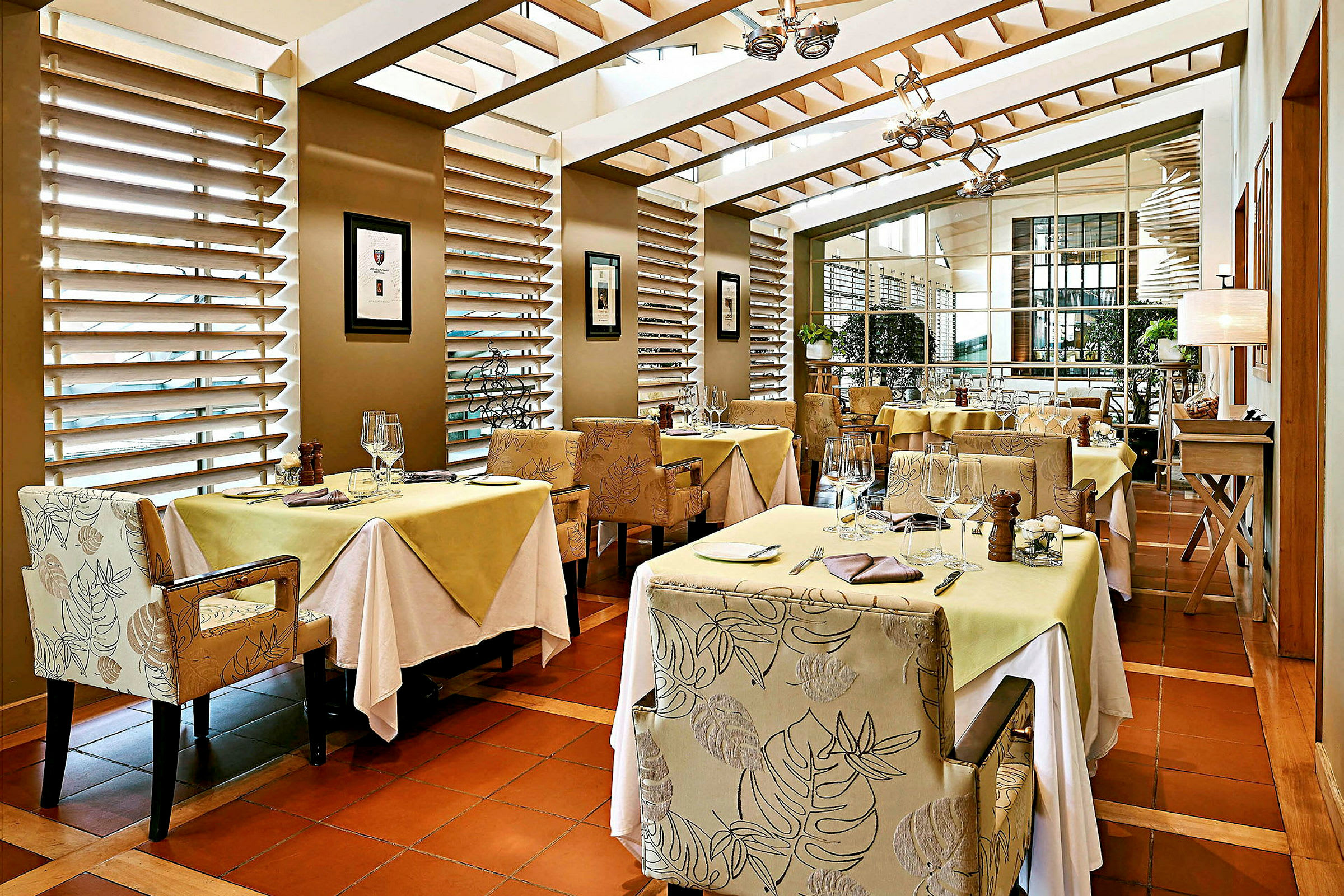 Et elegant restaurantområde med bord dekket for måltider, omgitt av stoler med mønstret stoff. Store vinduer slipper inn naturlig lys. (AI-generert tekst)