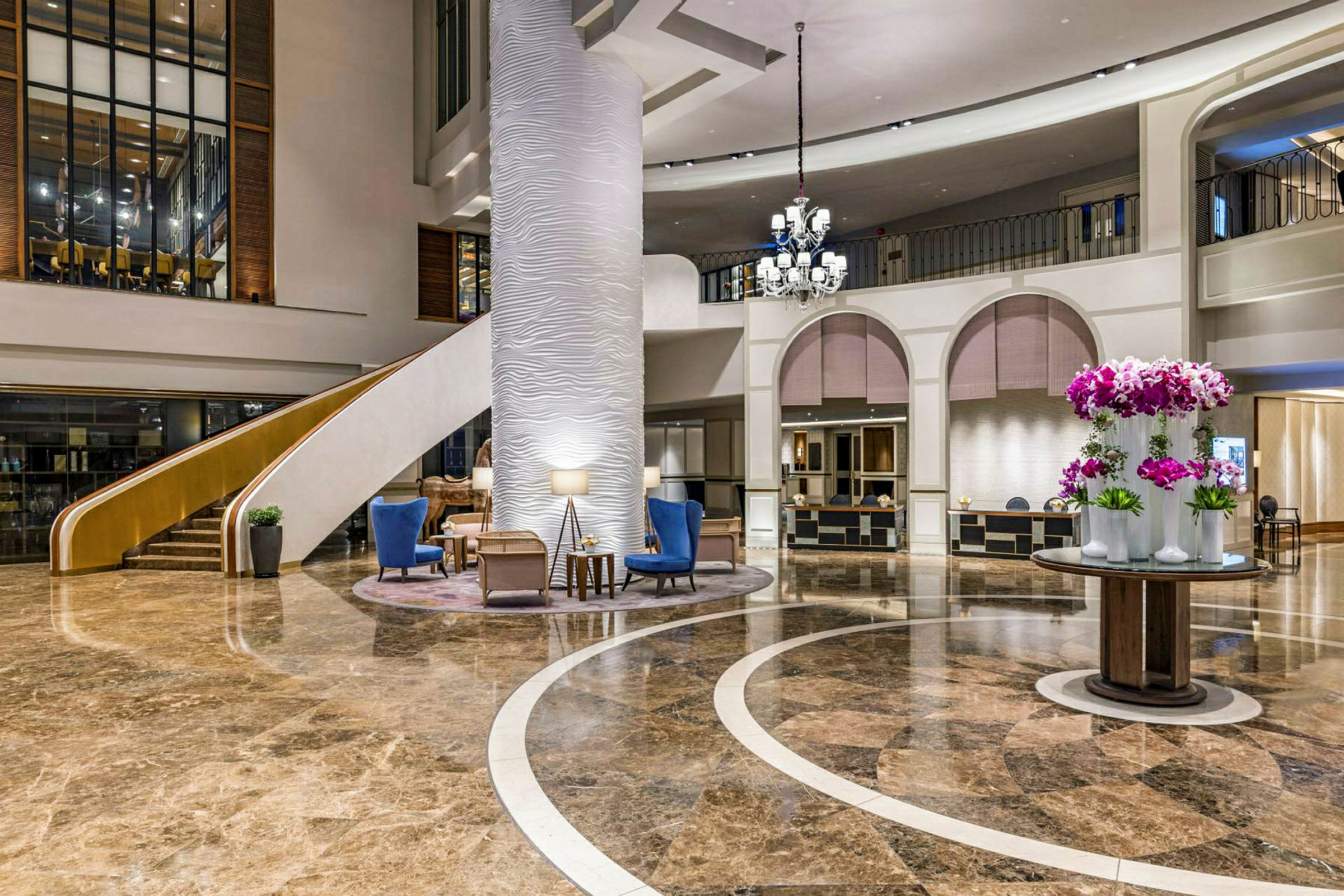 En hotellobby med marmorgulv, en stor søyle, en trapp, sittemøbler og en rund borddekorasjon med blomster. (AI-generert tekst)