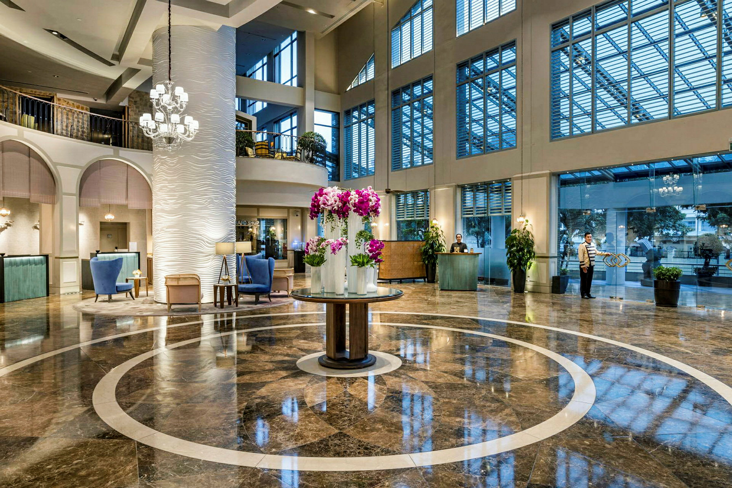 En elegant hotellobby med glanset marmorgulv og et sentralt rundt område dekorert med lilla blomster i vaser. Høye vinduer slipper inn lys, og en resepsjon sees i bakgrunnen. (AI-generert tekst)