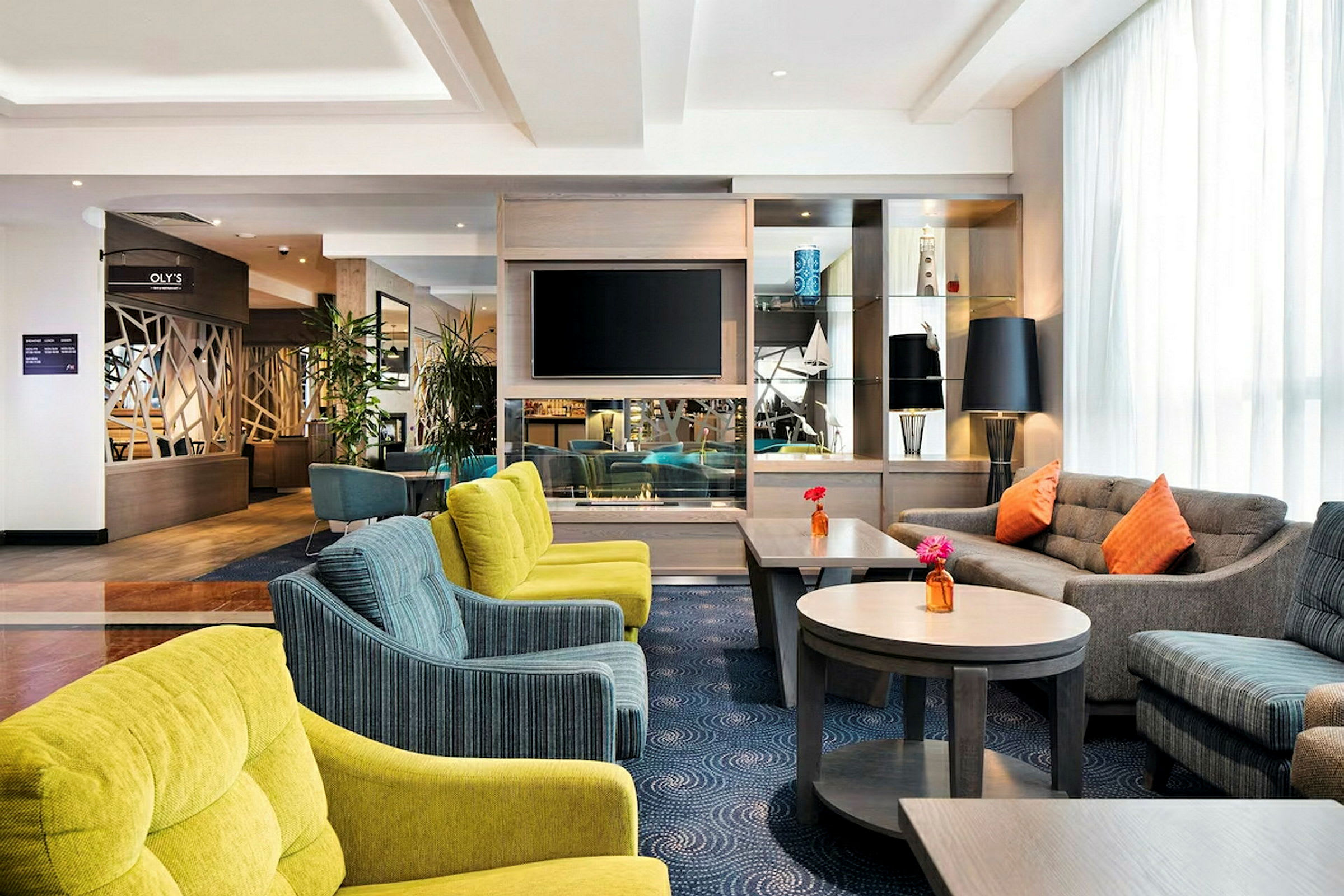 Et hotellobby med komfortable sitteplasser, flere sofaer og stoler i ulike farger, samt bord med blomster. En TV er montert på veggen og det finnes dekorative gjenstander på hyllene. (AI-generert tekst)