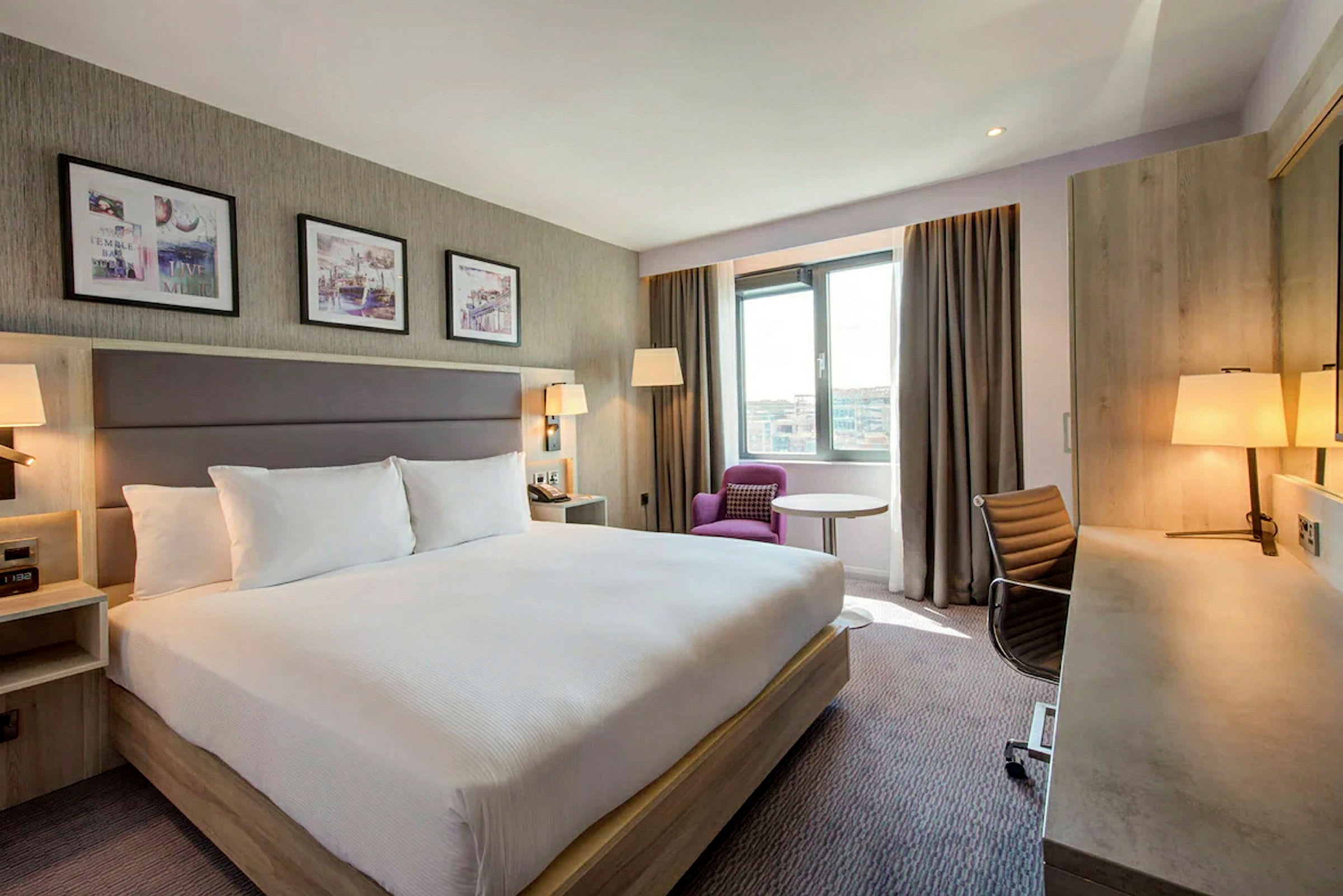 Et hotellrom med en stor dobbeltseng, skrivebord, stol og en lenestol ved vinduet. (AI-generert tekst)