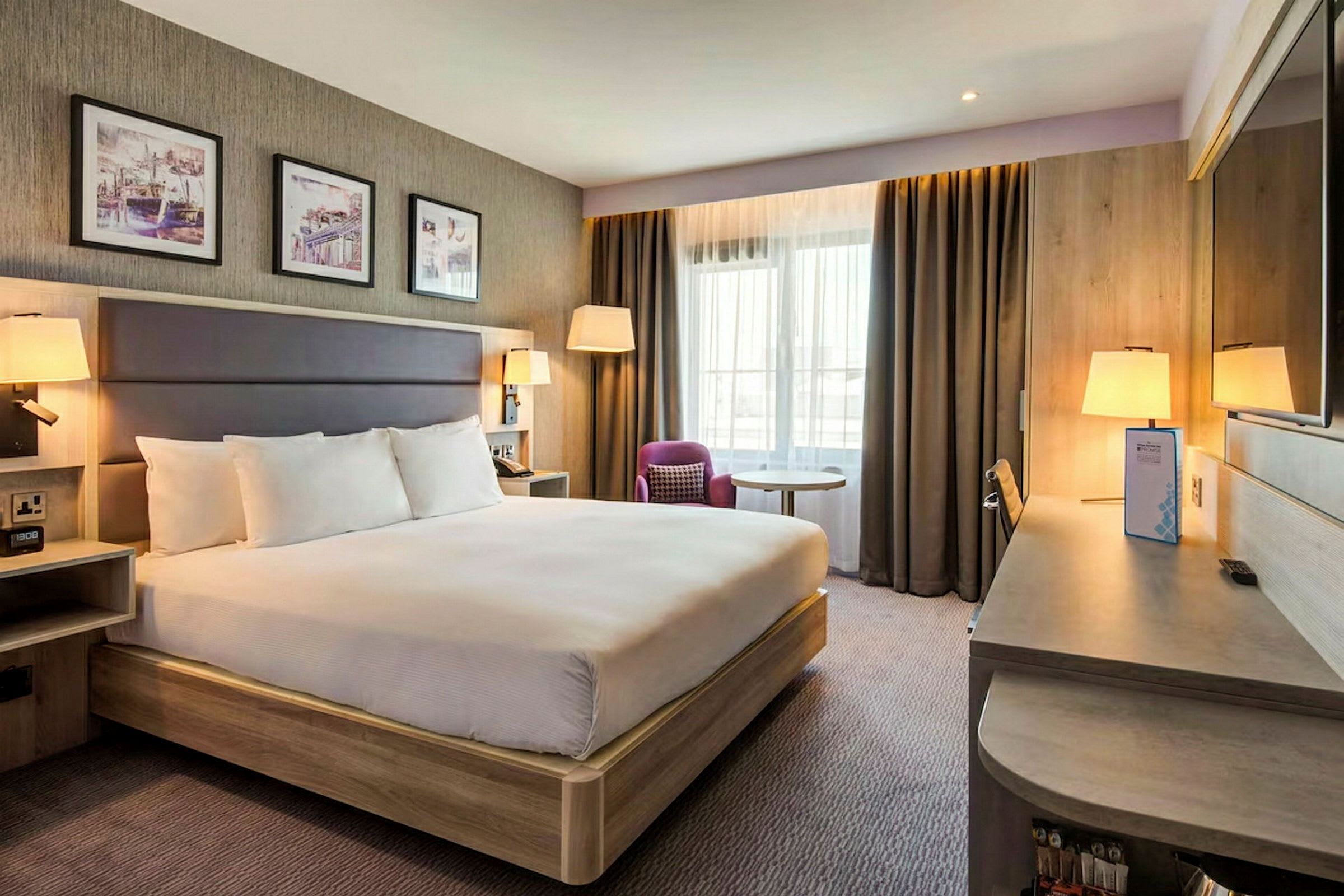 Hotellrom med en stor seng, skrivebord, stol og vindu med gardiner. (AI-generert tekst)