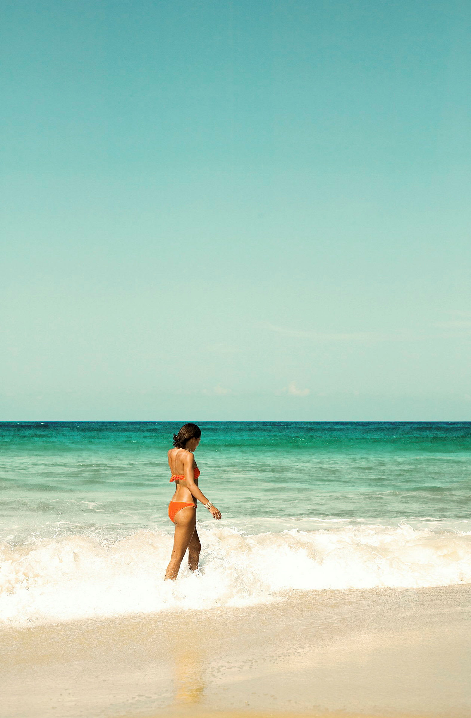 En kvinne i oransje bikini går ved stranden nær bølgene. (AI-generert tekst)