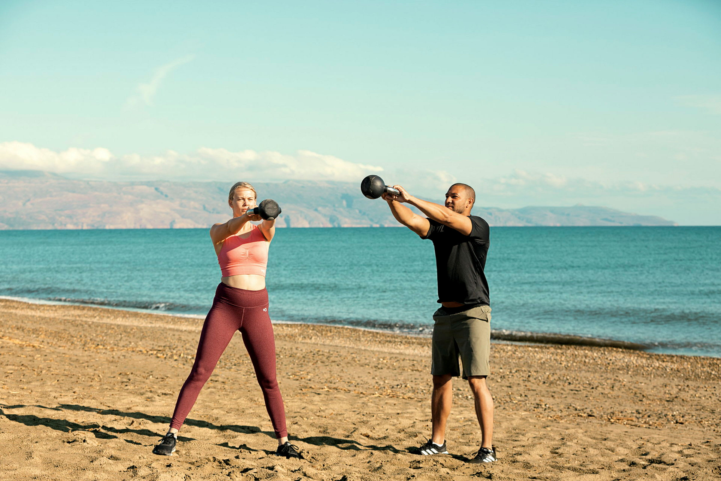 To personer trener med kettlebells på en sandstrand med havet i bakgrunnen. (AI-generert tekst)