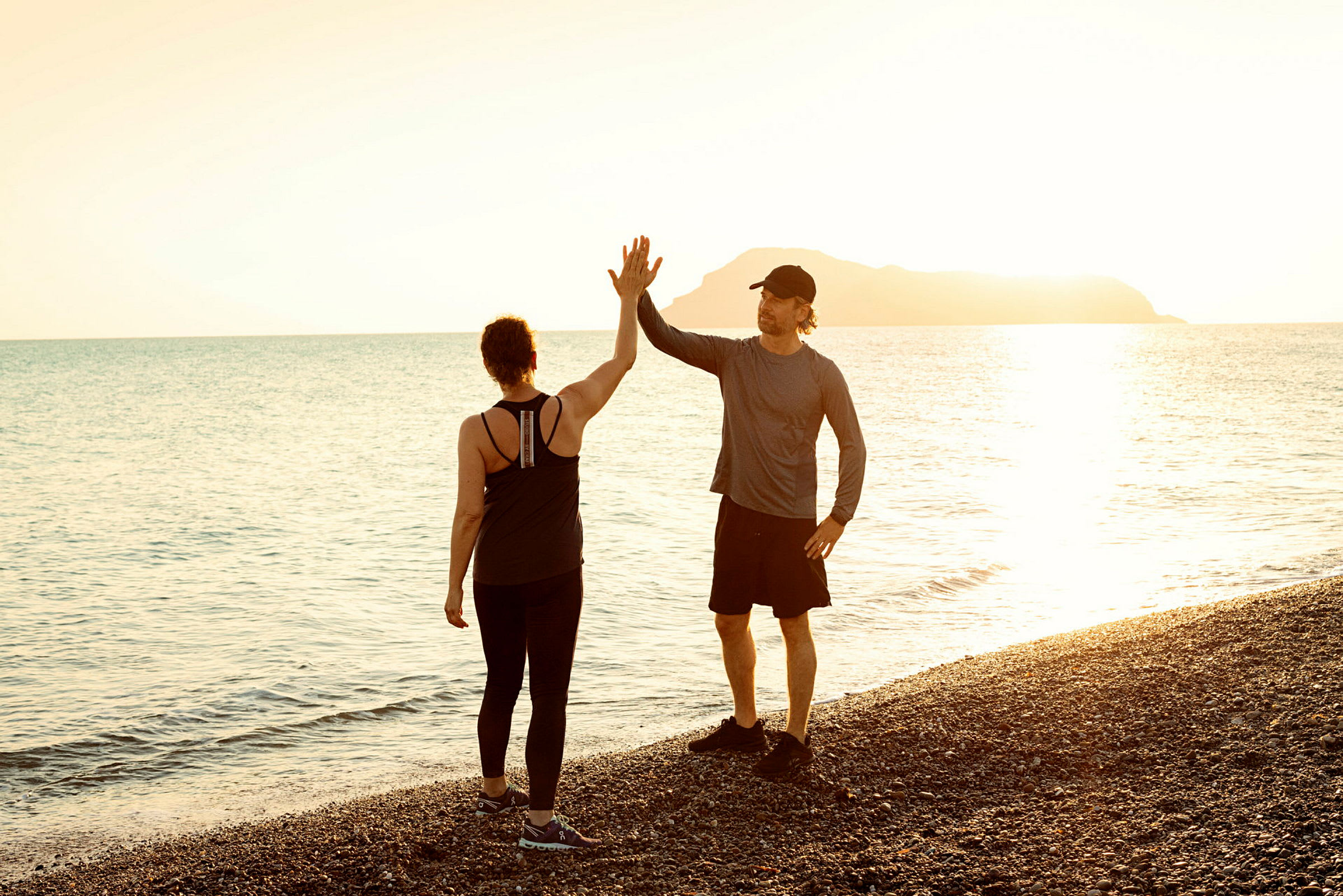 To personer står på en steinstrand og gir hverandre en high five under solnedgangen over havet. (AI-generert tekst)