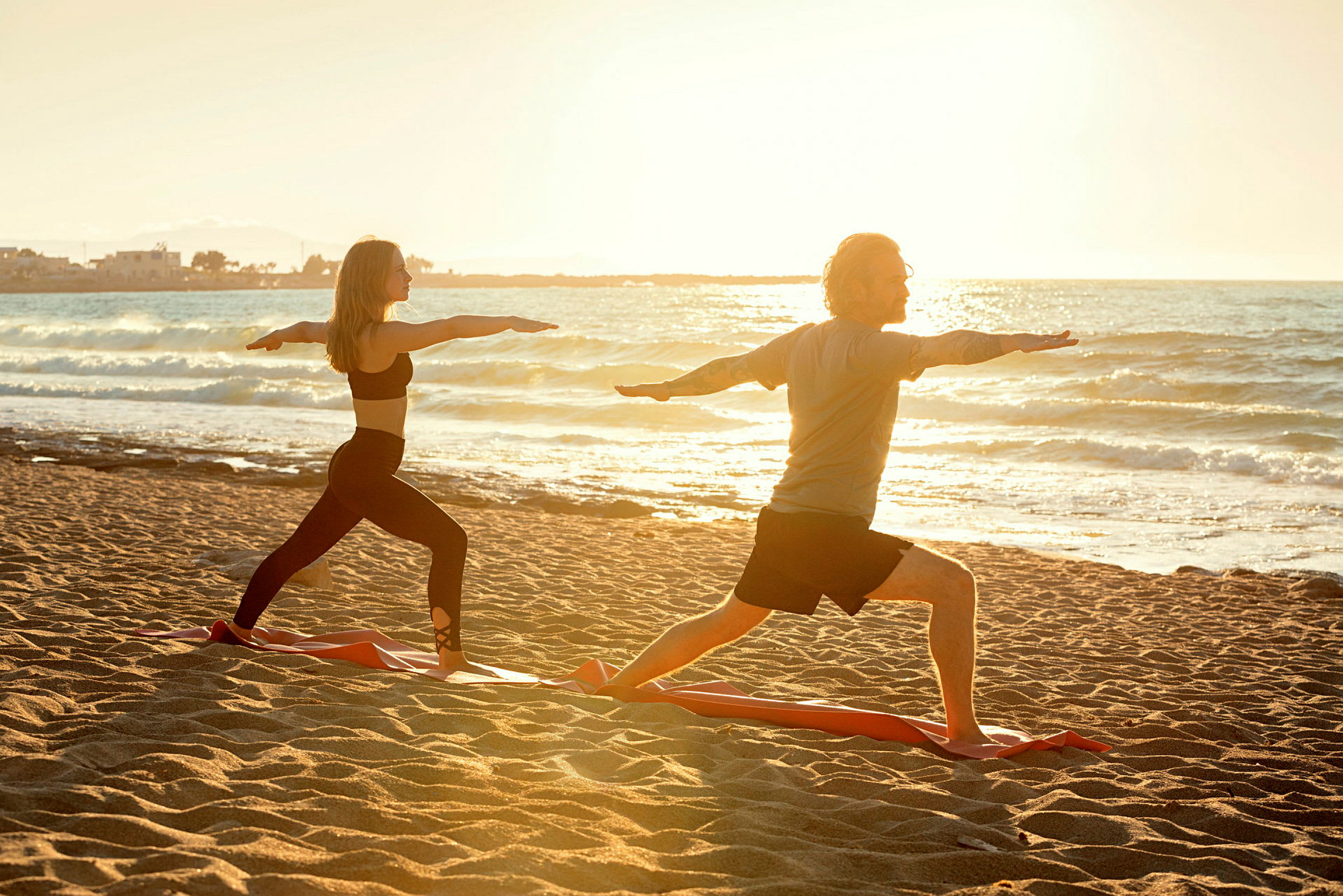 To personer utfører yogaøvelser på en sandstrand ved havet, med solen som går ned i det fjerne. (AI-generert tekst)