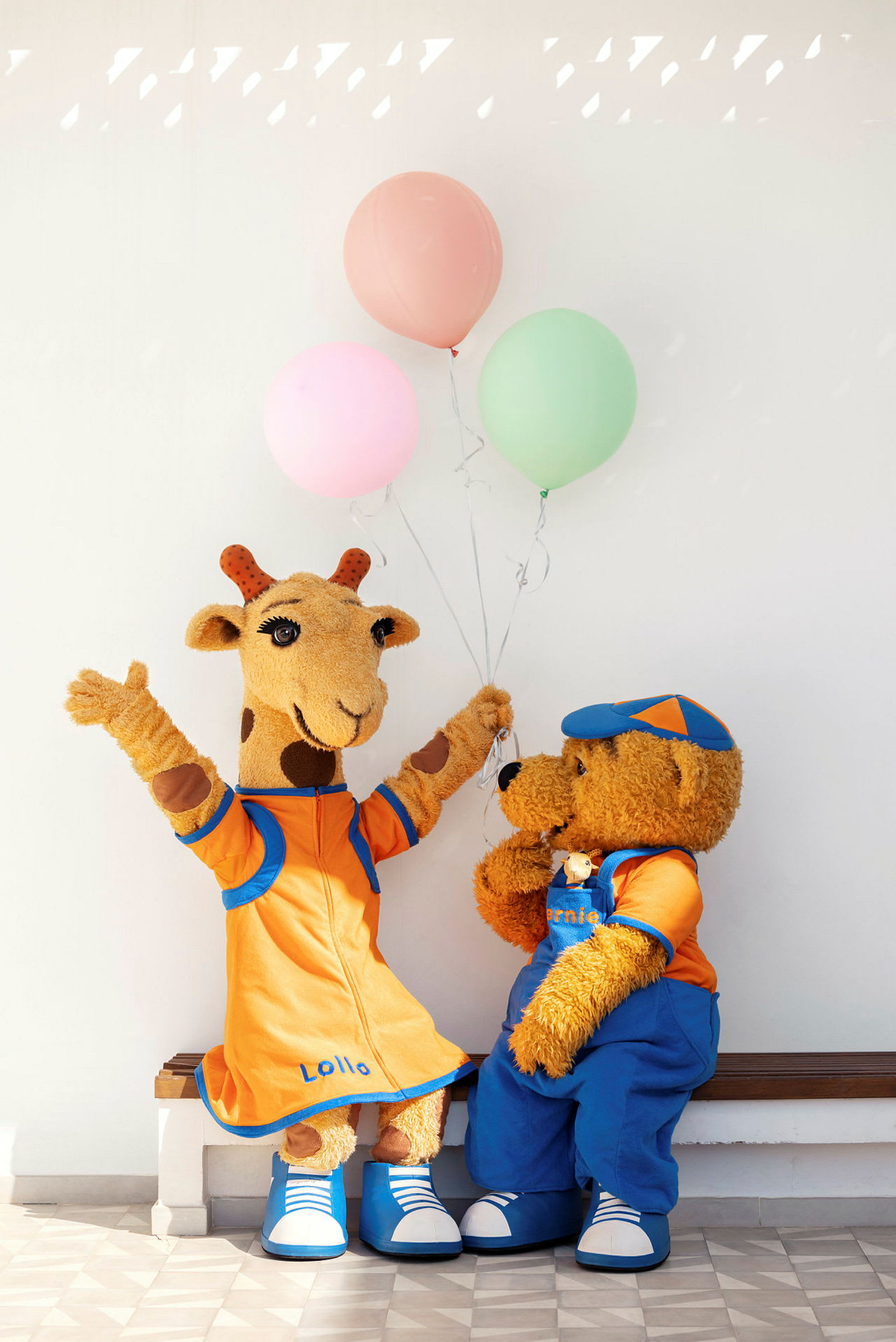 To maskoter, en giraff og en bjørn, sitter på en benk med ballonger. (AI-generert tekst)