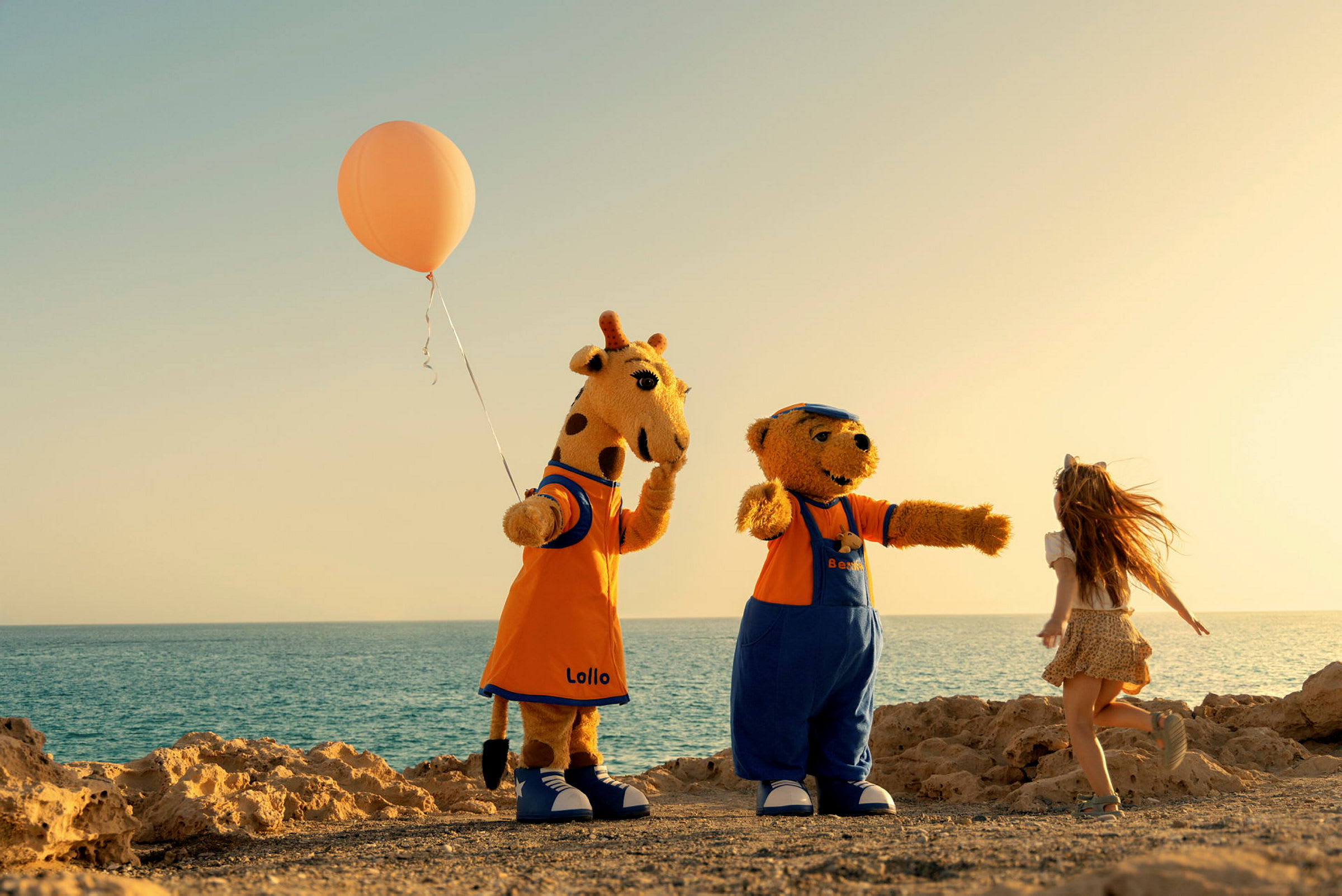 To figurer kledd som en giraff og en bjørn står ved siden av et barn på en strand med havet i bakgrunnen. Giraffen holder en oransje ballong. (AI-generert tekst)