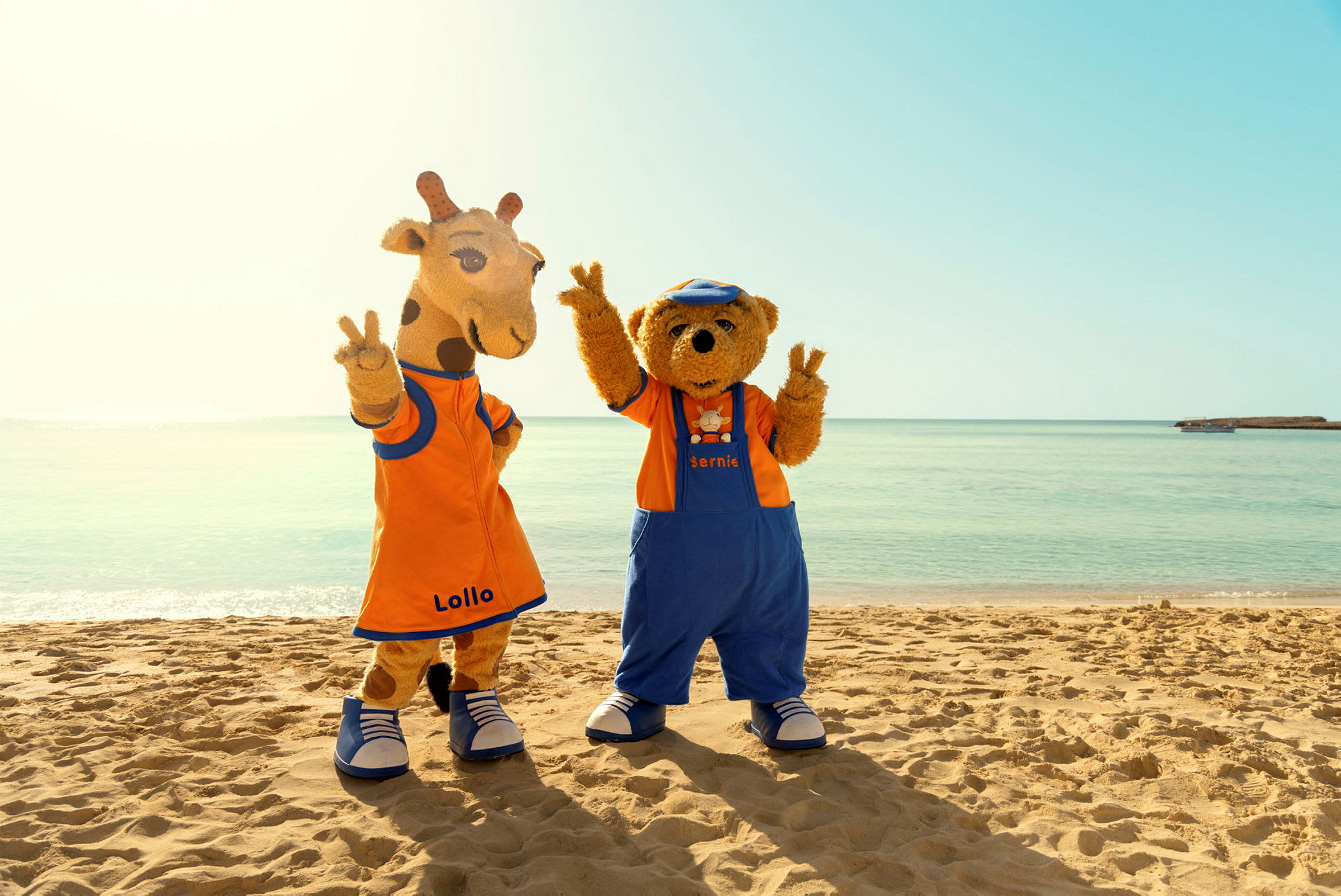 To maskotkarakterer med navnene Lollo og Bernie står på en strand foran det klare havet. (AI-generert tekst)