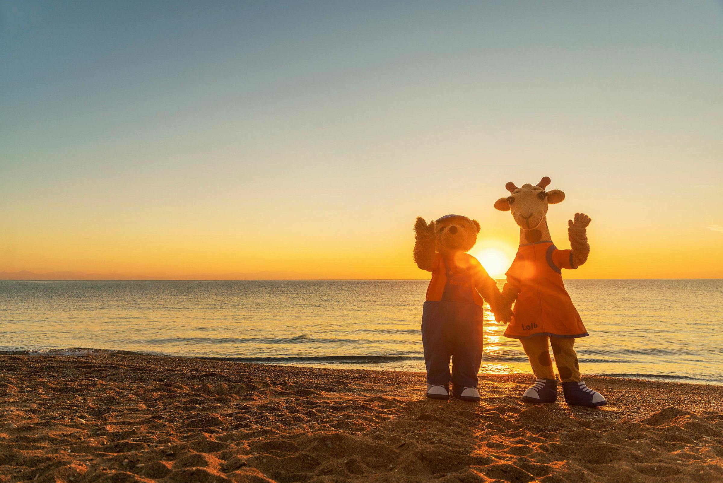 To maskoter står på en strand foran en solnedgang, vendt mot kameraet. (AI-generert tekst)